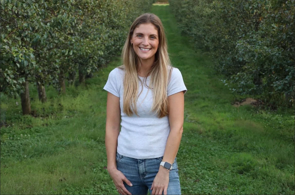A woman standing on a grassy path surrounded by bushes, smiling at the camera.