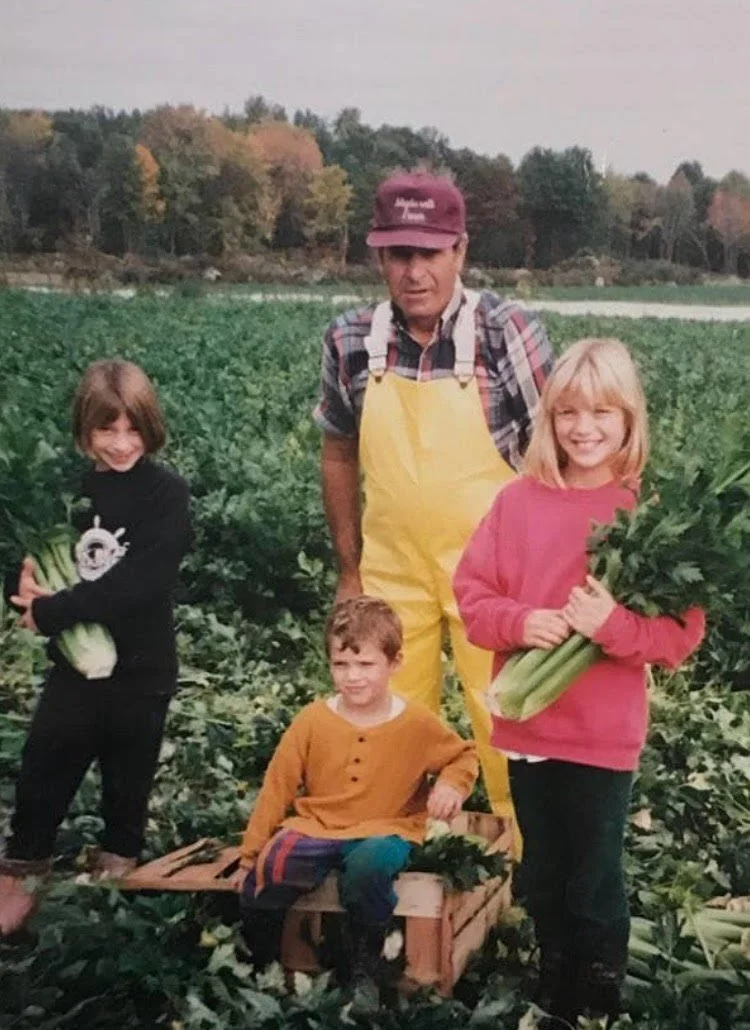 A group of four children and one adult standing and sitting in a lush green fruit or vegetable field, holding bunches of vegetables, with fall foliage in the background.
