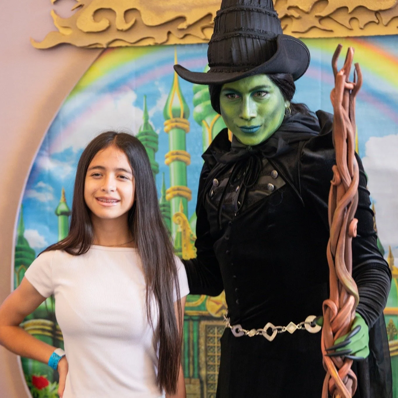 A girl with long brown hair smiling and standing next to a person dressed as the Wicked Witch of the West from The Wizard of Oz. The witch has green face paint, a tall black hat, and is holding a twisted staff. The background features a colorful, cartoonish castle and rainbow with blue skies.