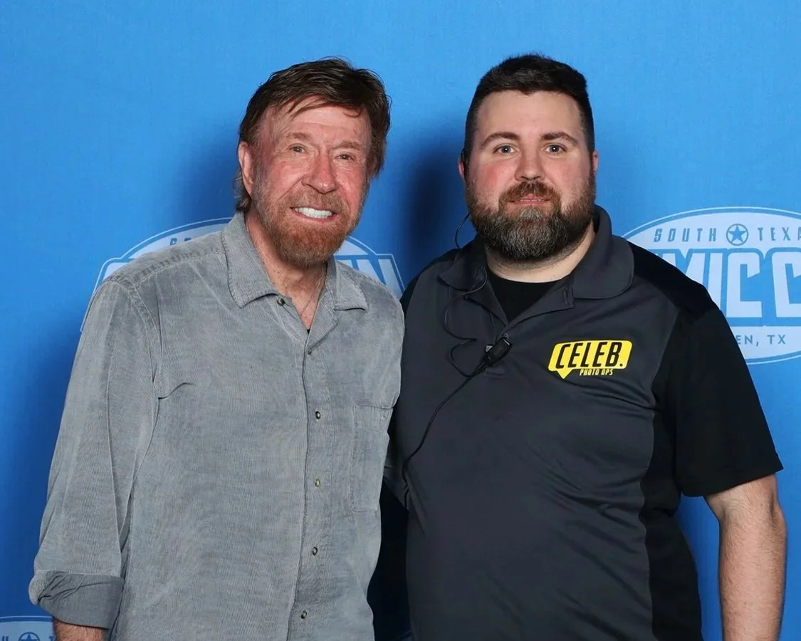 Two men posing together at an event in front of a blue backdrop with the words "South Texas" and "Comic" visible. The man on the left has medium-length brown hair and a beard, wearing a gray button-up shirt. The man on the right has short dark hair, a full beard, and is wearing a black shirt with a yellow patch that says "CELEB" on his chest.