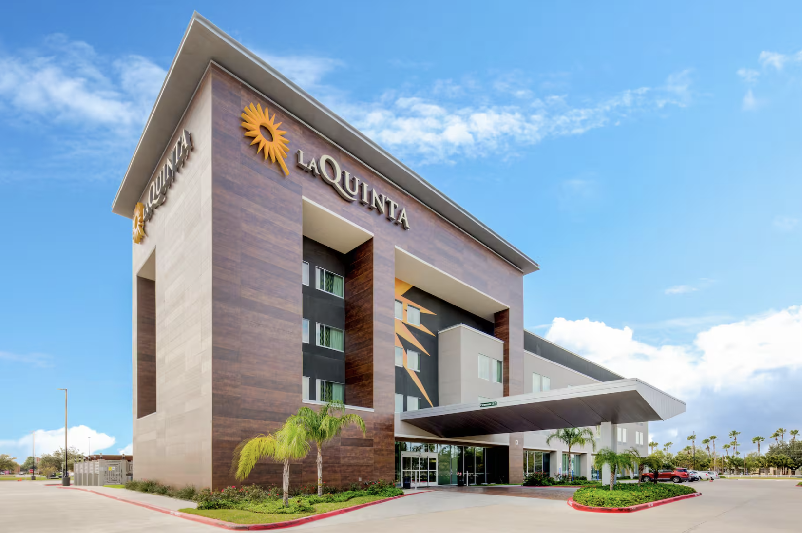 Exterior of a modern hotel building with the name La Quinta, featuring a large awning over the entrance, decorative palm trees, and a parking lot under a blue sky with scattered clouds.