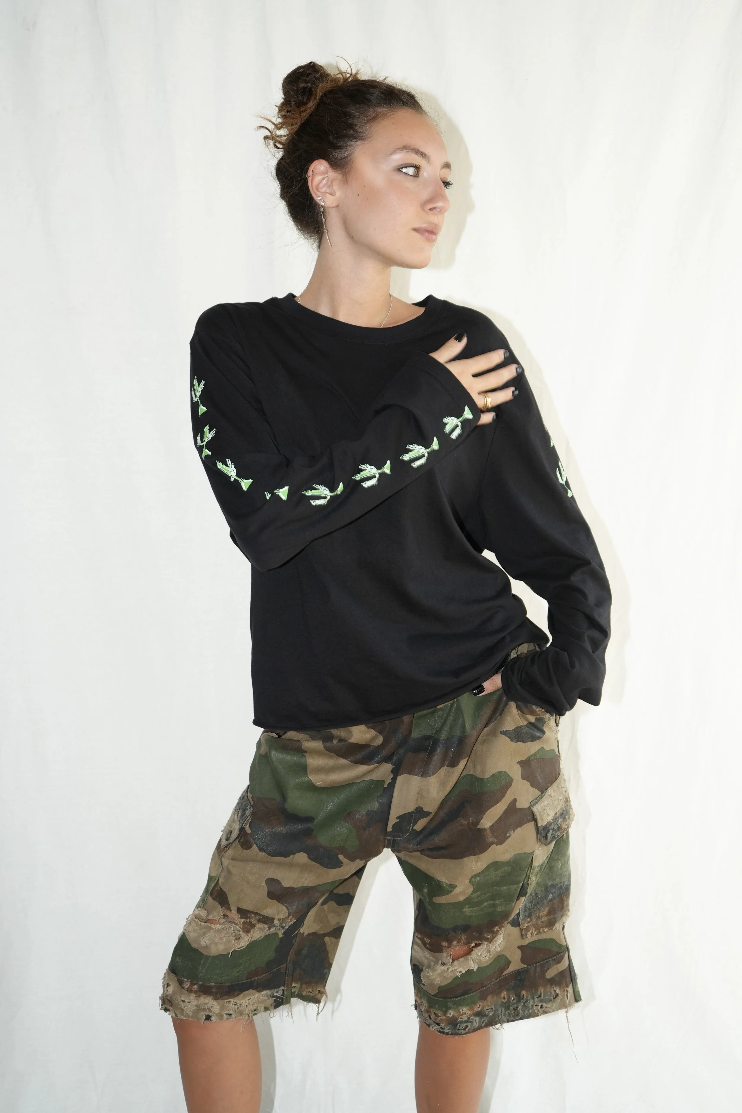 Une jeune femme, les cheveux attachés, porte un sweat-shirt noir à coutures décoratives sur les manches et un short camouflage, posant une main sur l'épaule sur fond blanc.