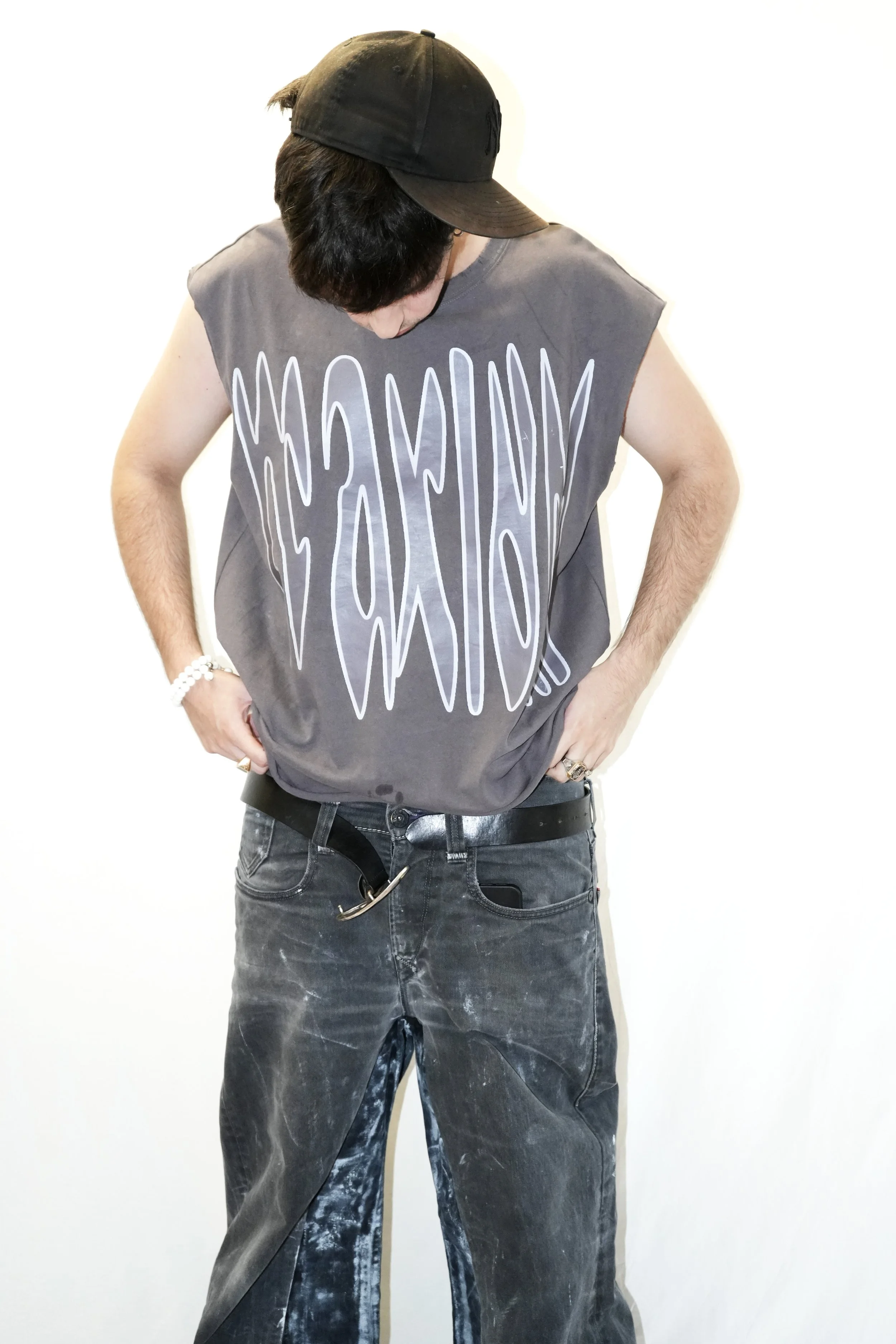 Une personne portant un chapeau noir, un débardeur gris à grandes inscriptions et un pantalon noir délavé, la tête baissée et les mains sur les hanches.