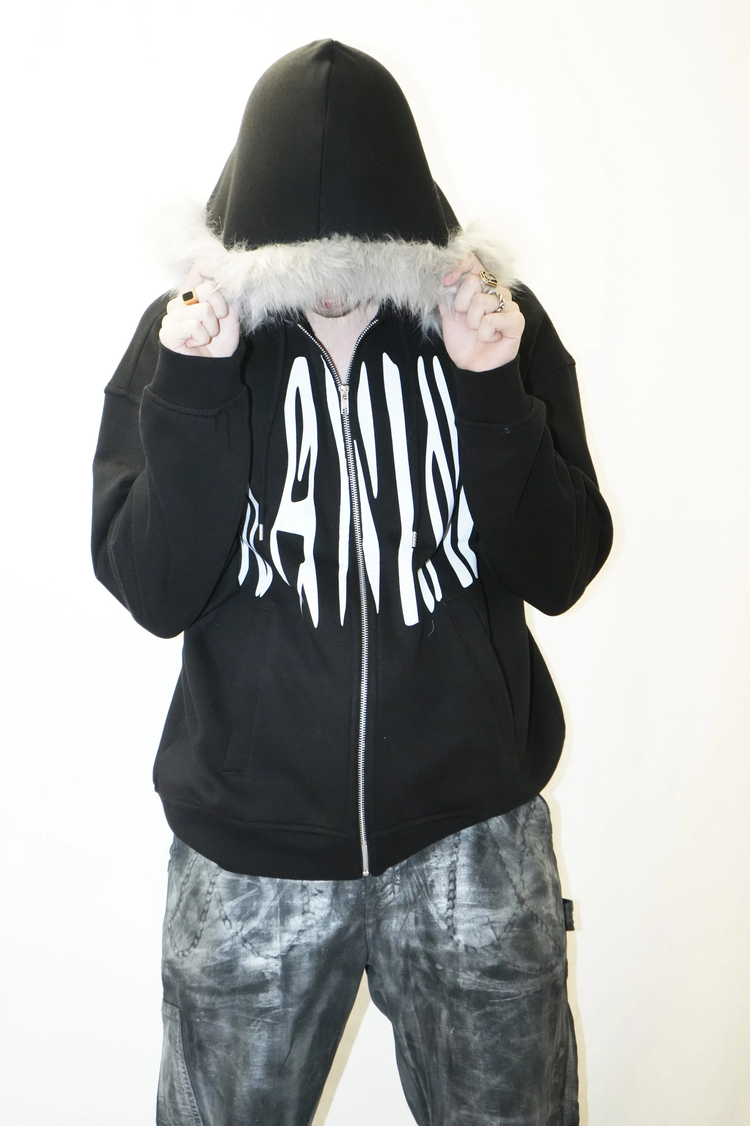 Une personne portant un sweat à capuche noir zippé avec de la fourrure sur la capuche, maintient la capuche rabattue sur son visage