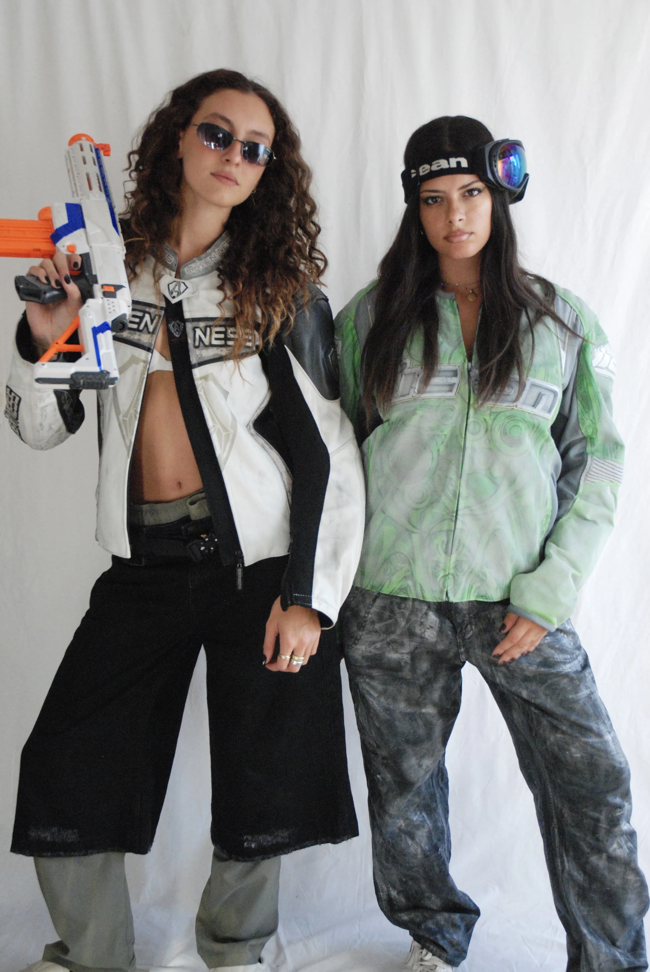 Deux femmes déguisées en personnages de jeux vidéo, l'une avec un pistolet jouet, portant des vêtements de sport et des lunettes de soleil, posent devant un fond blanc.