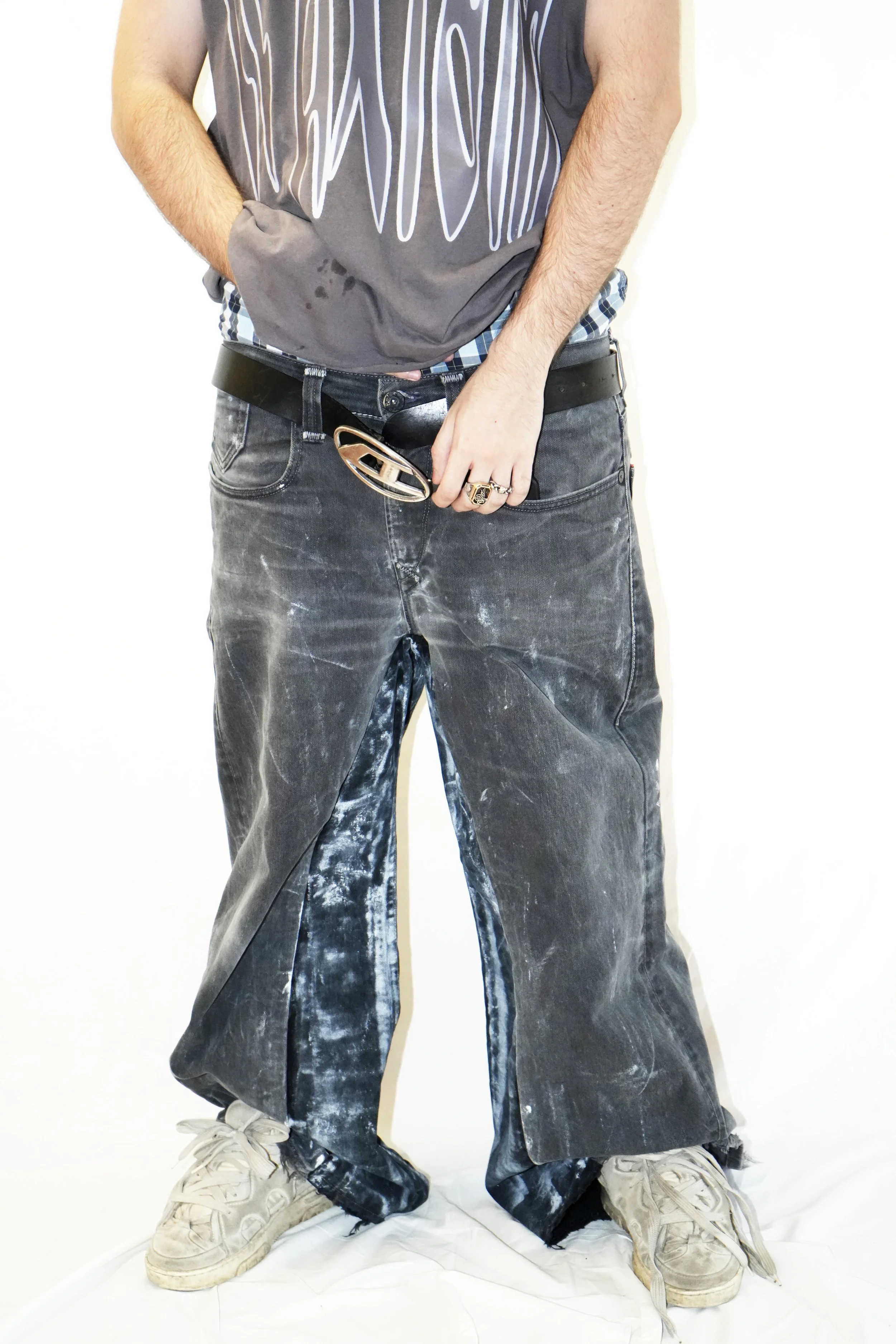 Personne portant un t-shirt gris, un pantalon ample délavé et des chaussures blanches usées, la main au niveau de l'aine.