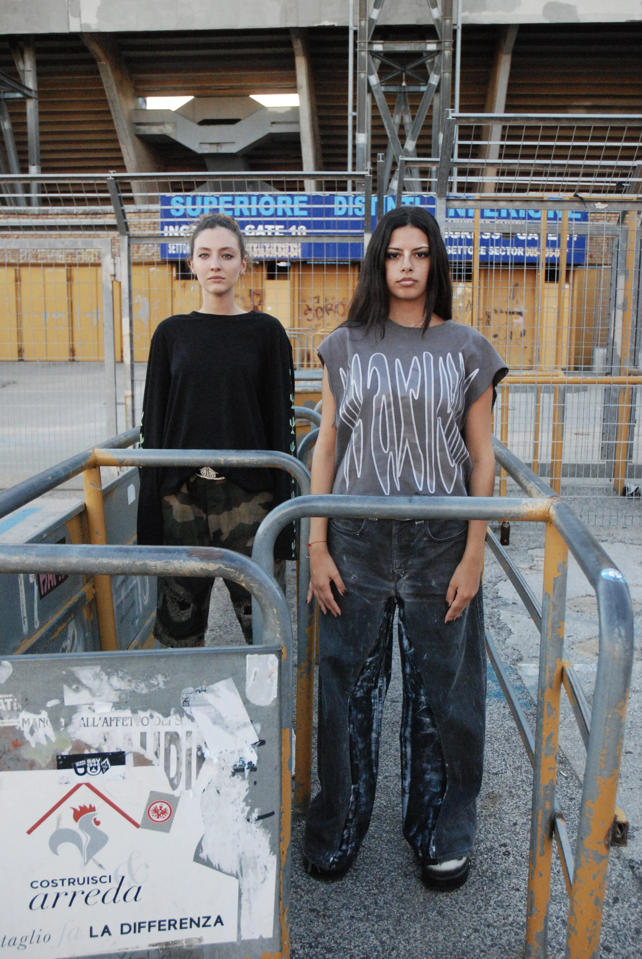 Deux jeunes femmes se tiennent debout dans un chantier, entourées de barrières métalliques. À l'arrière-plan, un panneau d'affichage bleu et une structure en béton se dressent au-dessus d'une zone clôturée.