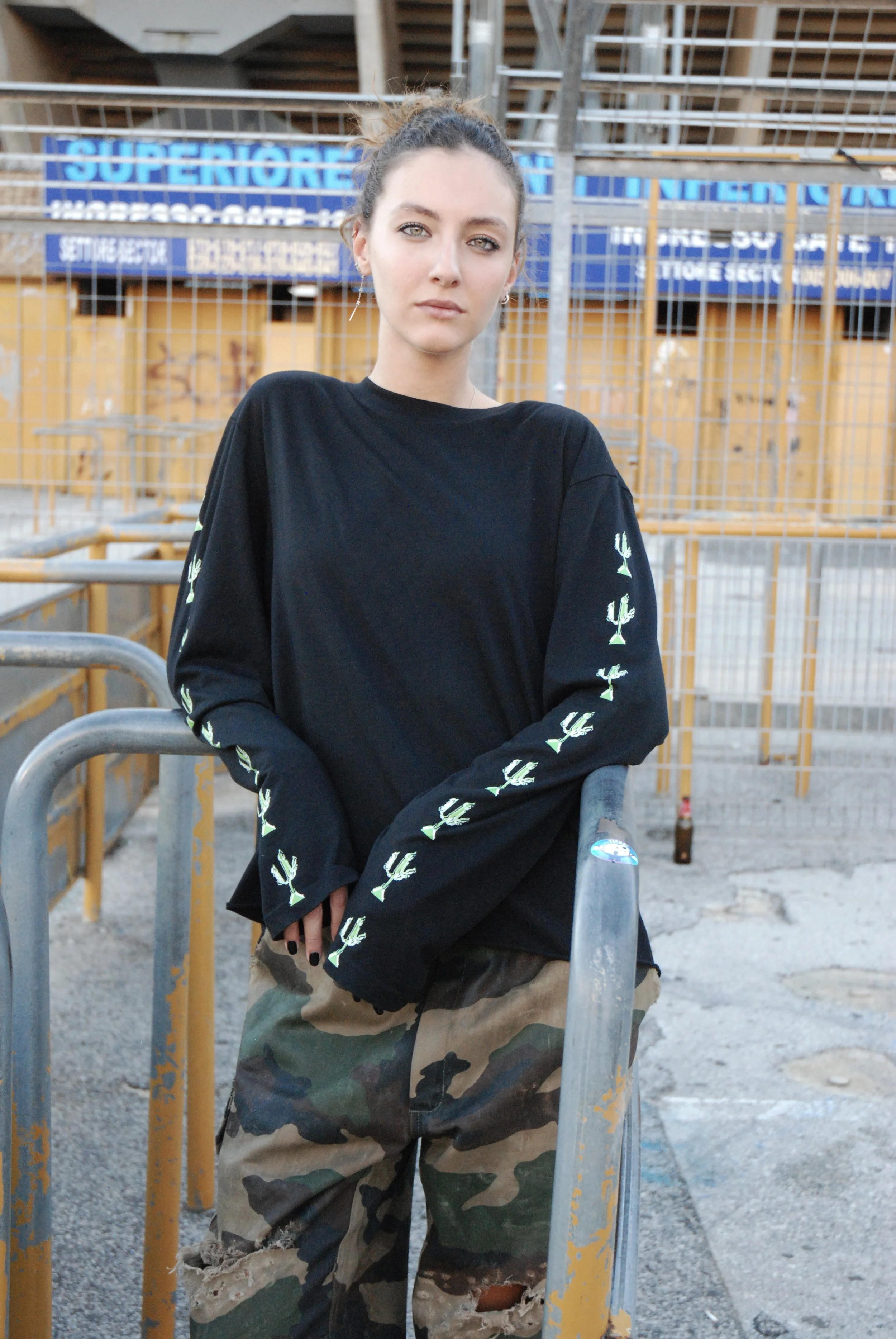 Jeune fille portant un pull noir à motifs de tournevis sur les manches et un pantalon cargo militaire, debout devant une clôture métallique en zone urbaine.