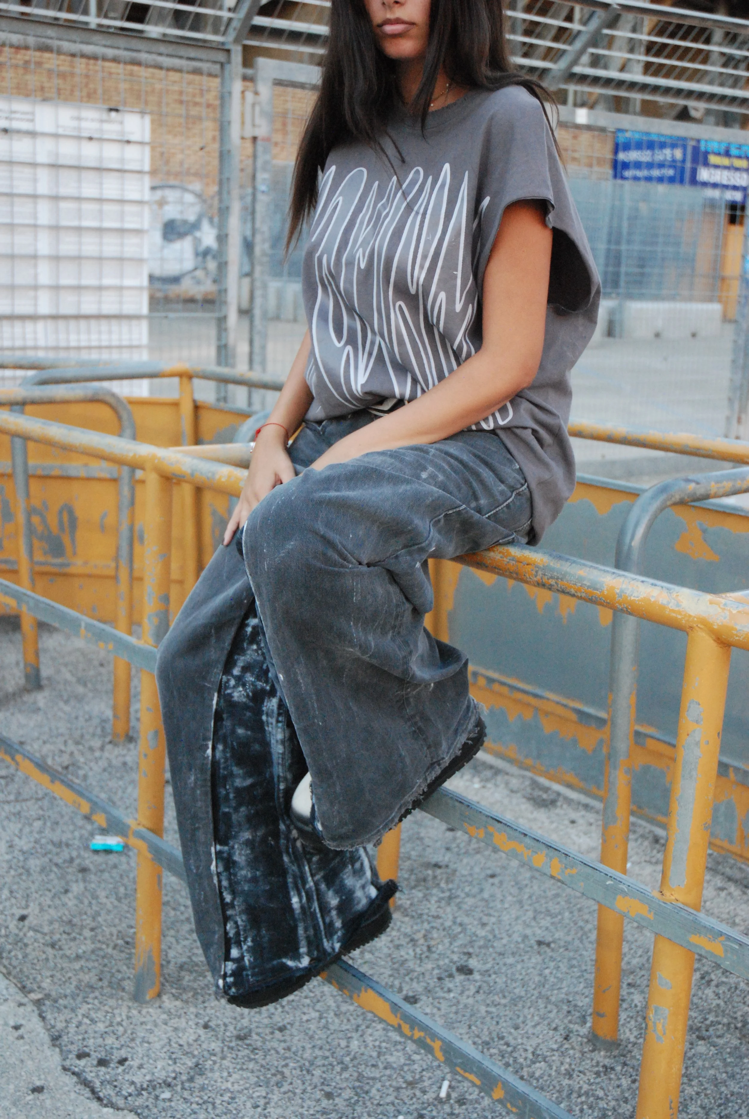 Une jeune fille assise sur une clôture métallique jaune, vêtue de vêtements décontractés, dans une zone industrielle extérieure.