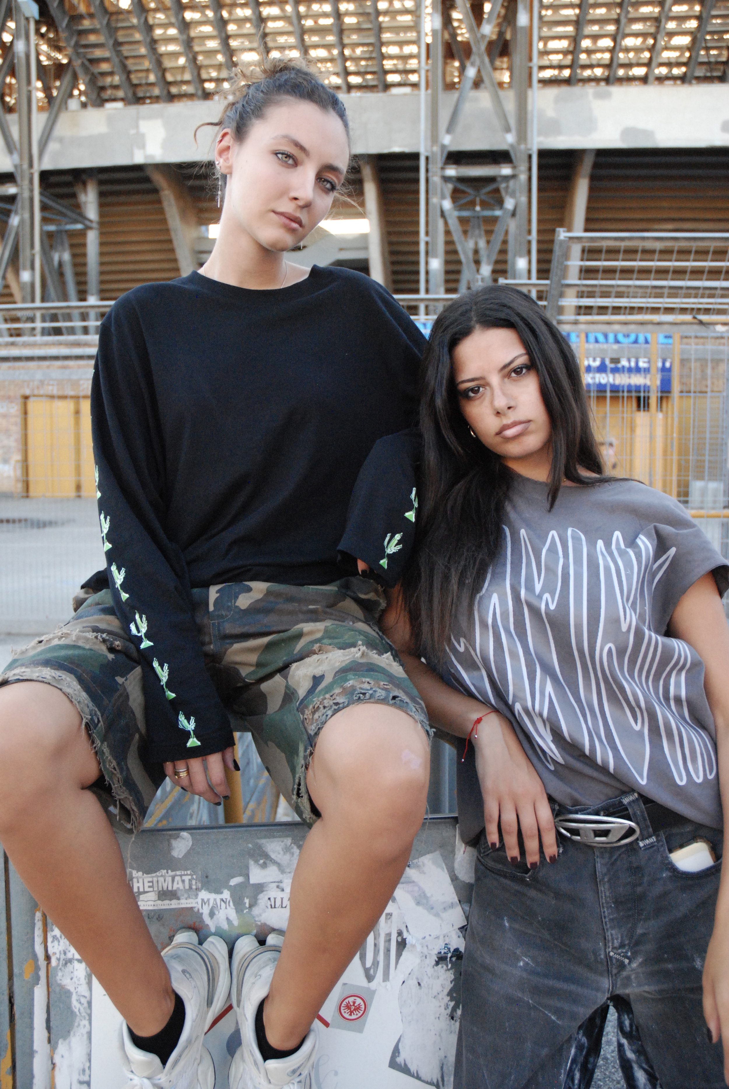 Deux jeunes femmes posent ensemble en extérieur, l'une en t-shirt noir et short camouflage, l'autre en t-shirt gris et jean noir.