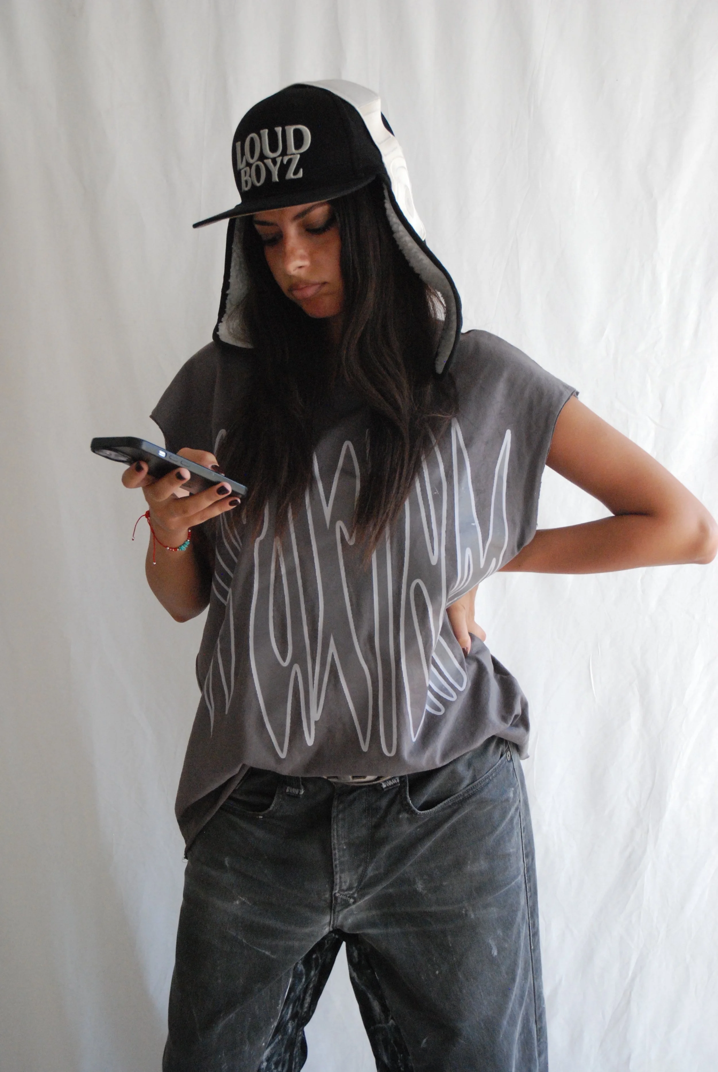 Une jeune fille portant une casquette noire LouD Boyz, un t-shirt gris avec des inscriptions noires et un jean noir sale, regarde son téléphone portable.