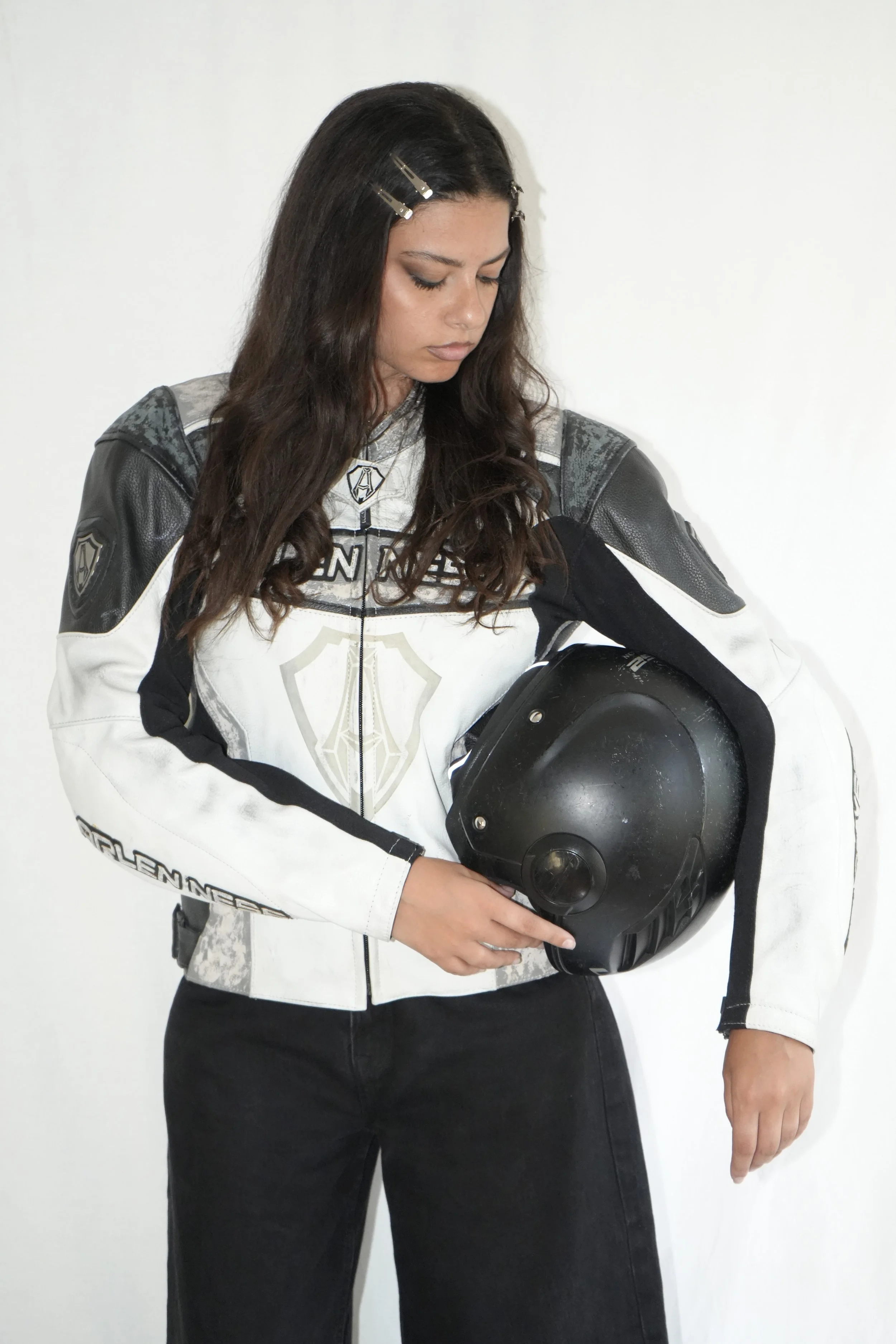 Femme en blouson de moto noir et blanc tenant un casque noir à la main, la tête légèrement inclinée vers le bas.