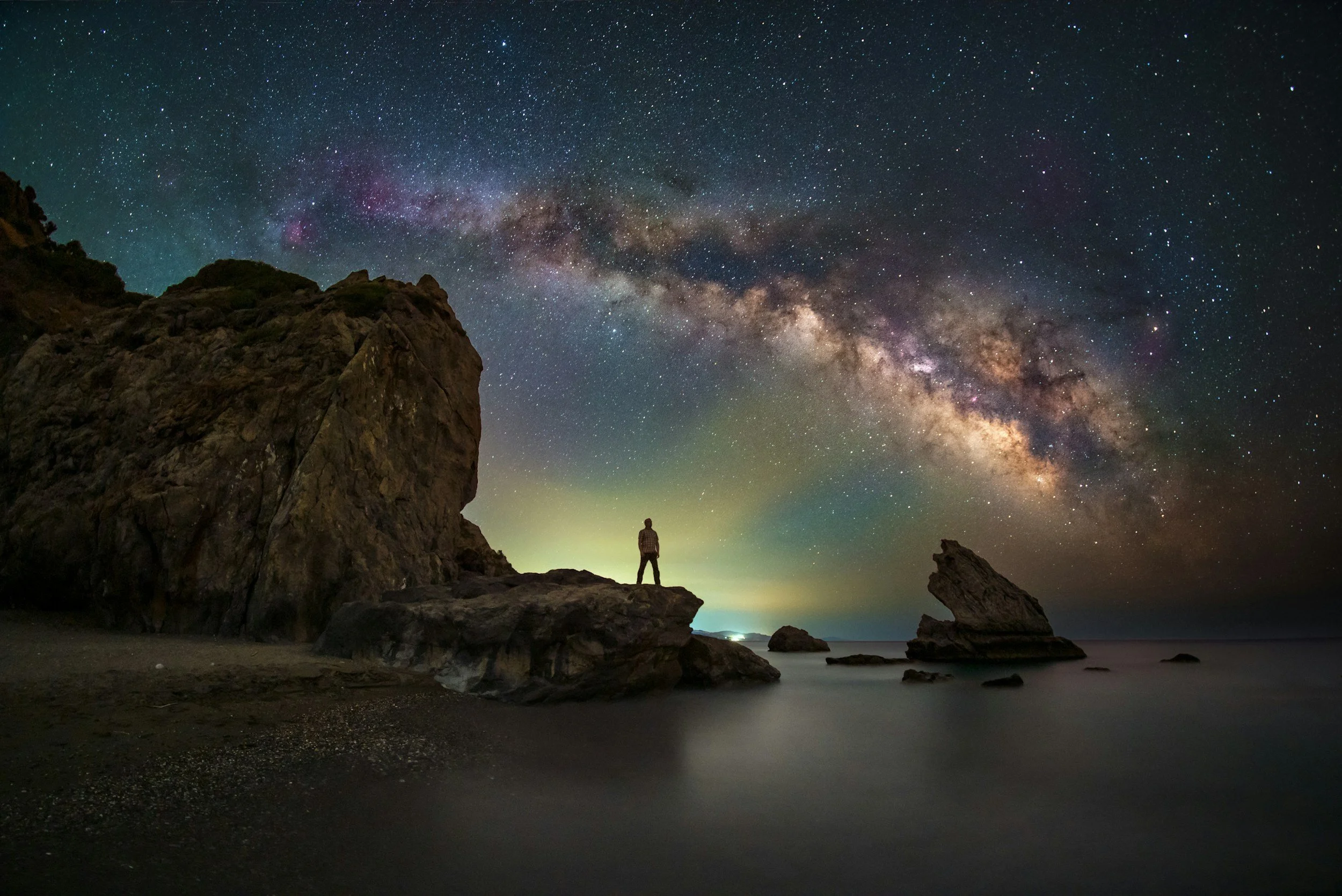 A person standing on a rock at the edge of the ocean, under a starry night sky with the Milky Way galaxy visible.