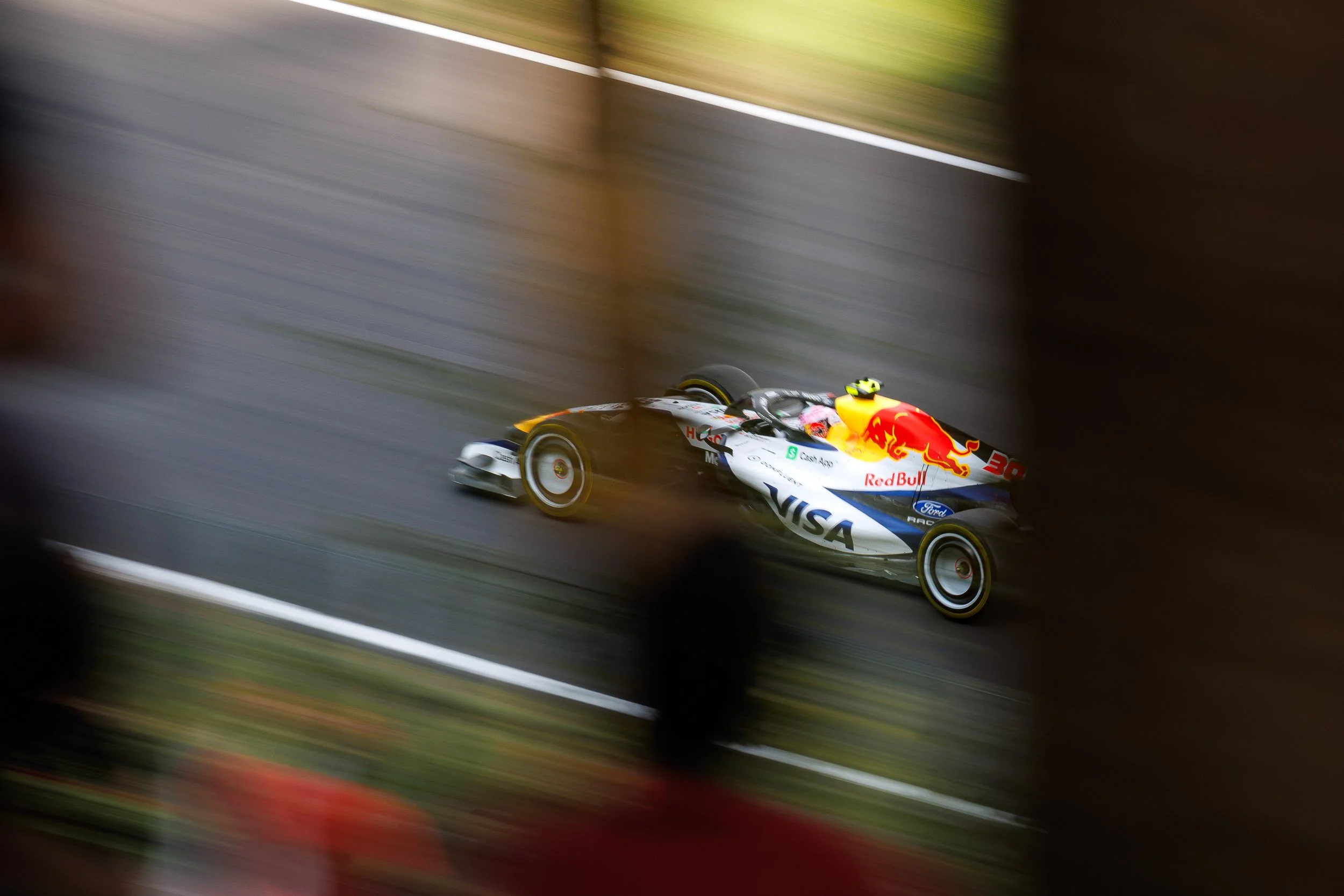 A race car with Red Bull sponsorship speeding along a racetrack, with motion blur indicating high speed, framed by safety barriers.
