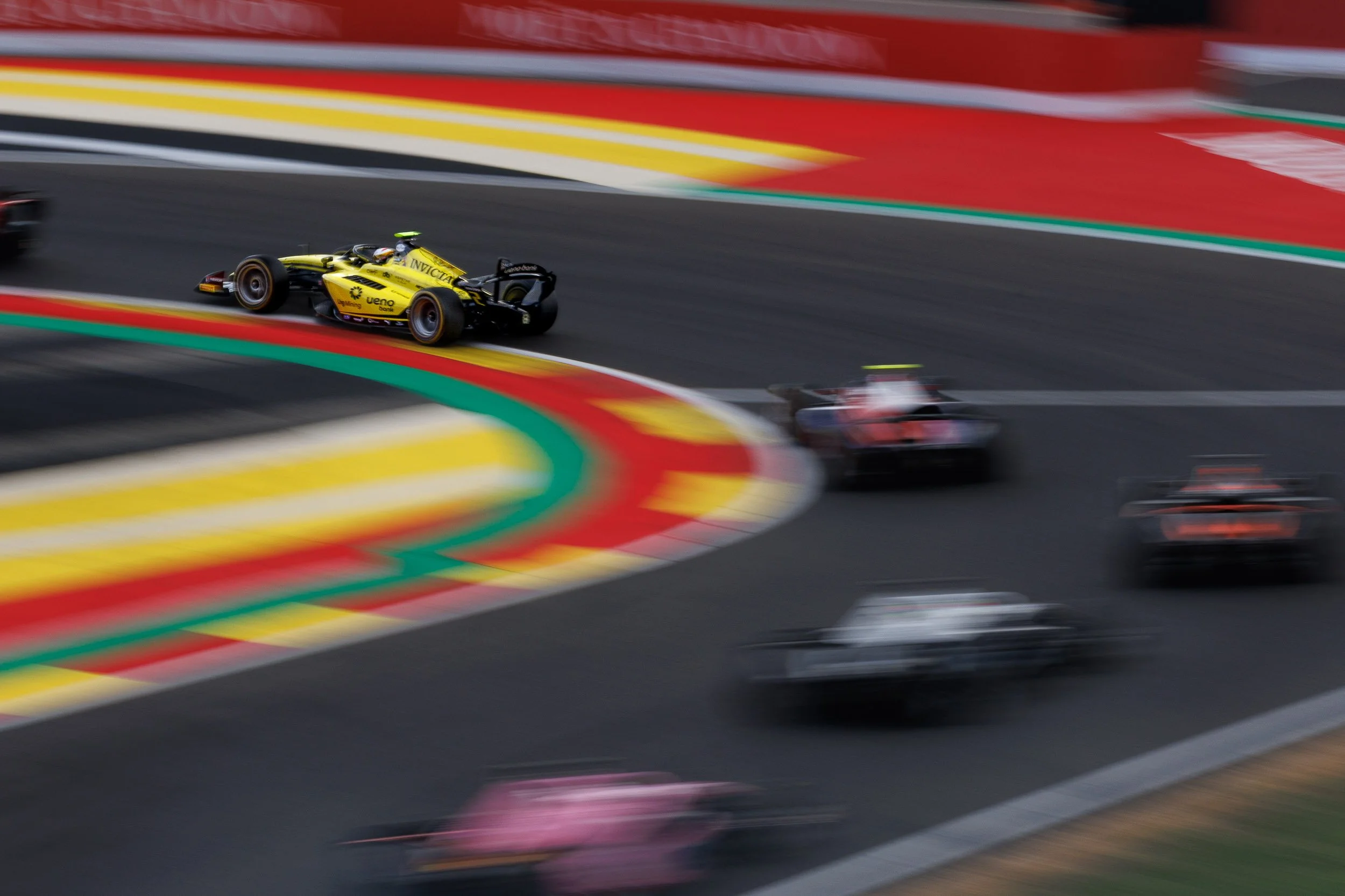 A yellow and black race car speeding on a racetrack with colorful curbing, blurred to show high speed, surrounded by other race cars.