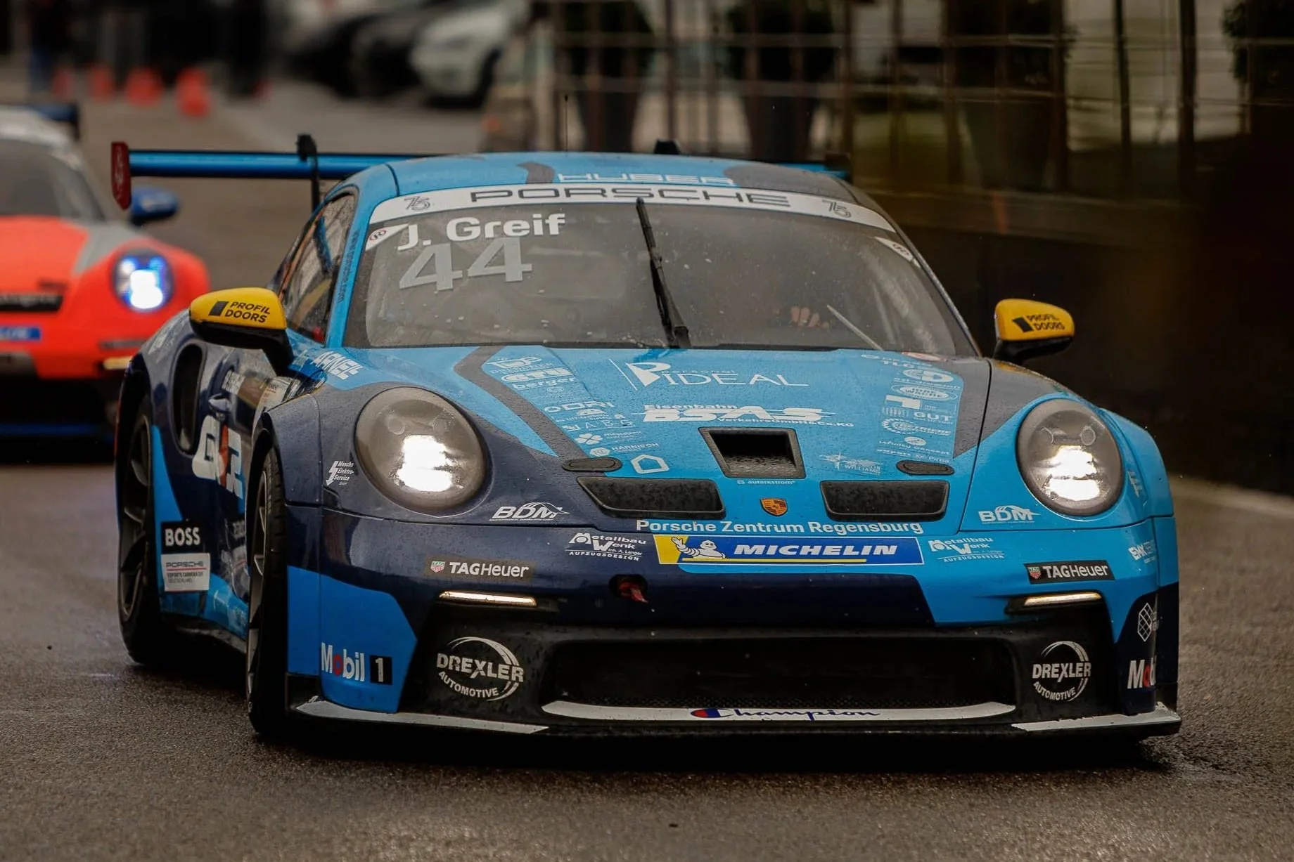 Blue Porsche race car with the number 44, driven by J. Greif, on a wet race track with a red car behind it and a blurred background.