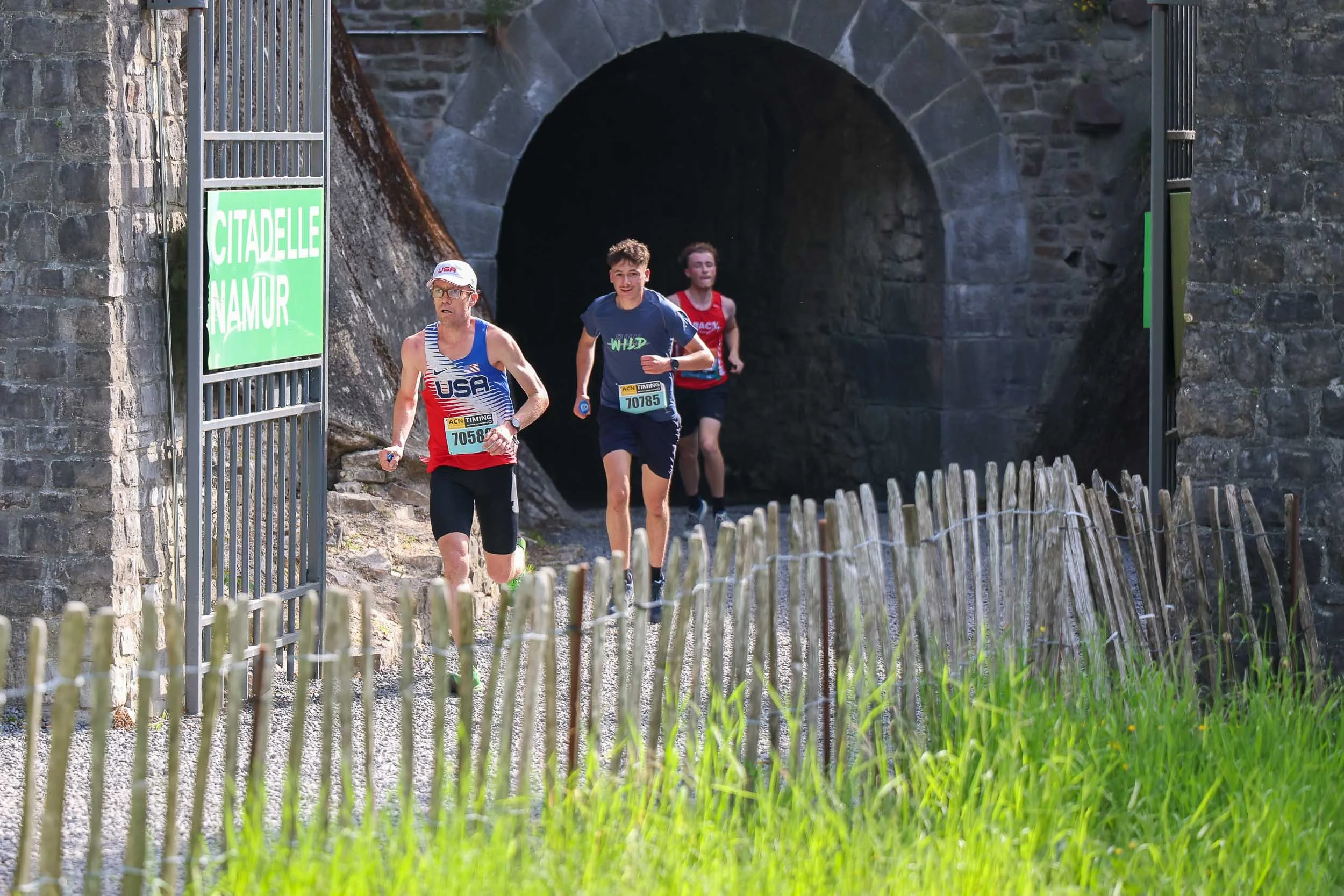Three runners participating in a race emerging from a tunnel under an old stone bridge during daytime.