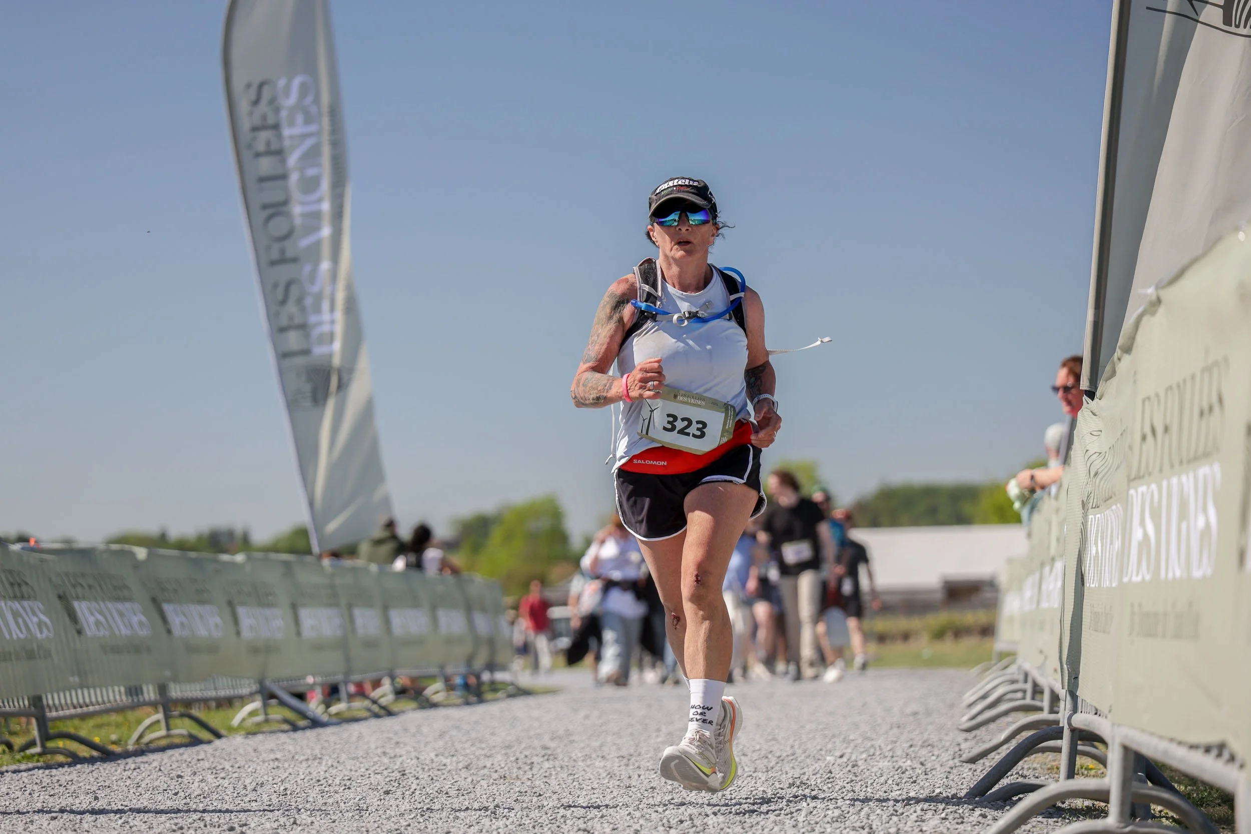 A female runner in a white tank top, black shorts, and sunglasses runs during a race, with a bib number 323, near a barrier with a blue sky in the background.