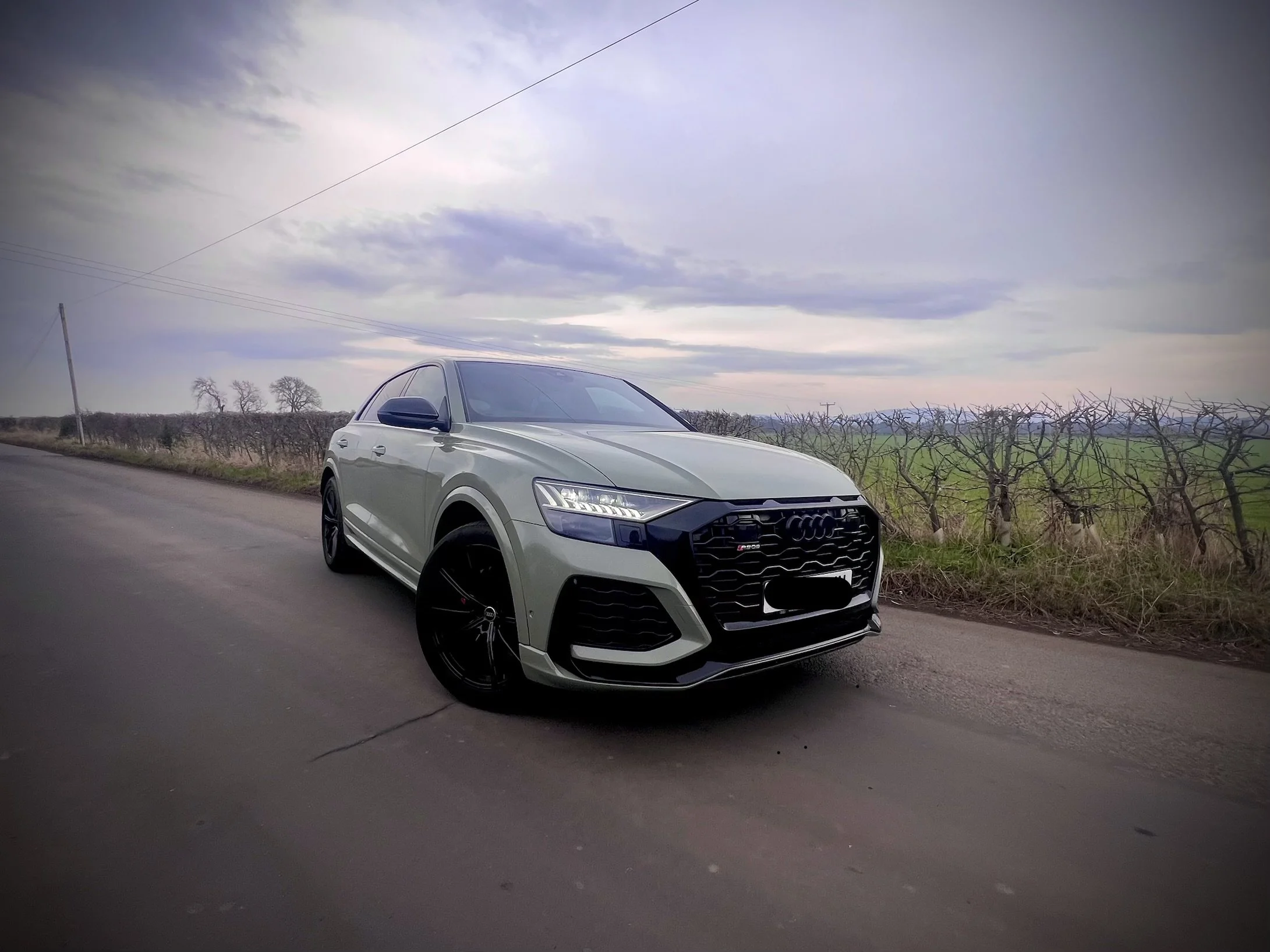 A silver Audi SUV parked on the side of a rural road with leafless trees and overcast sky in the background.