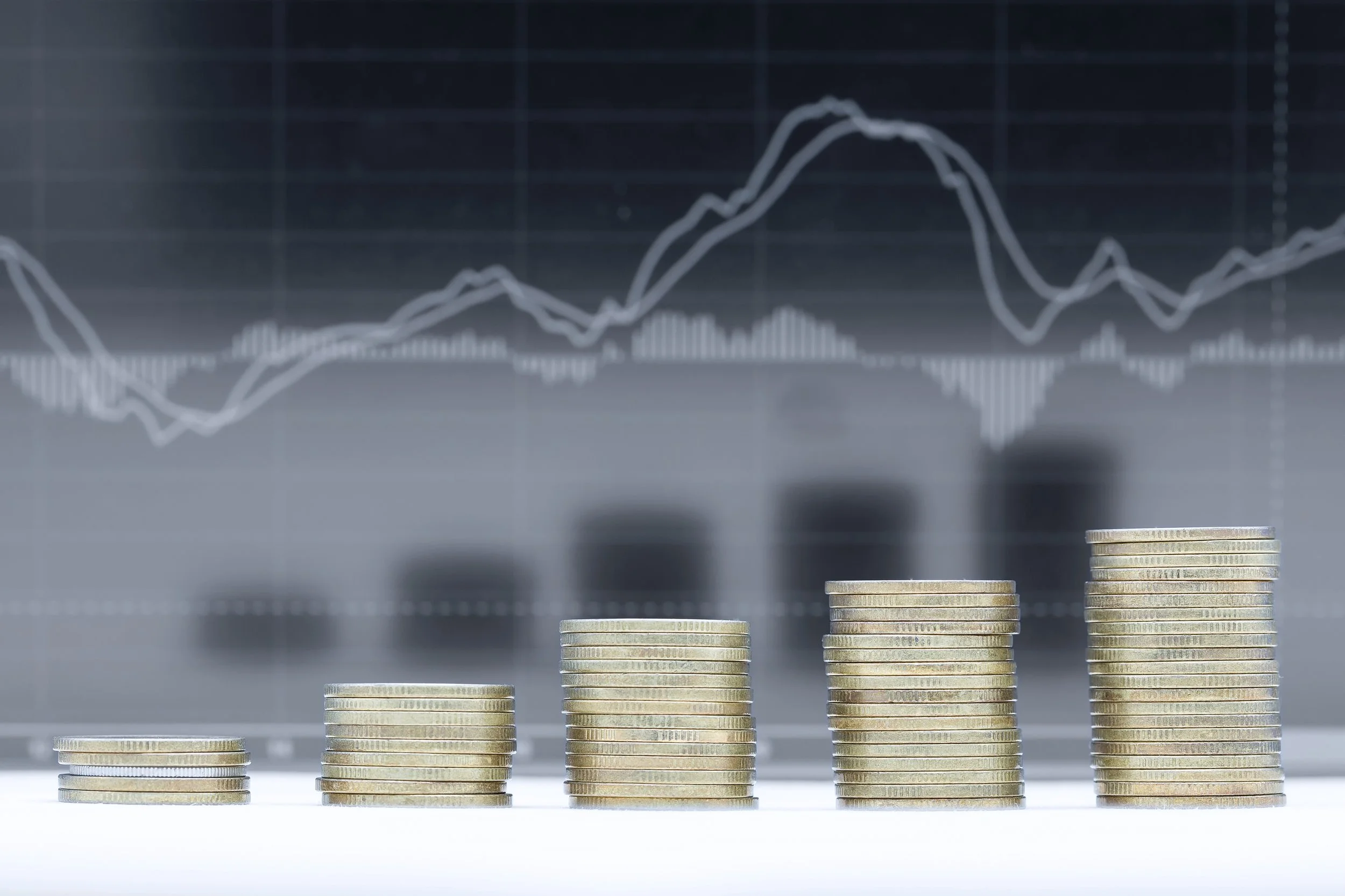 Stacks of coins progressing in height from left to right, with a blurred financial chart in the background showing line graphs and bar charts.