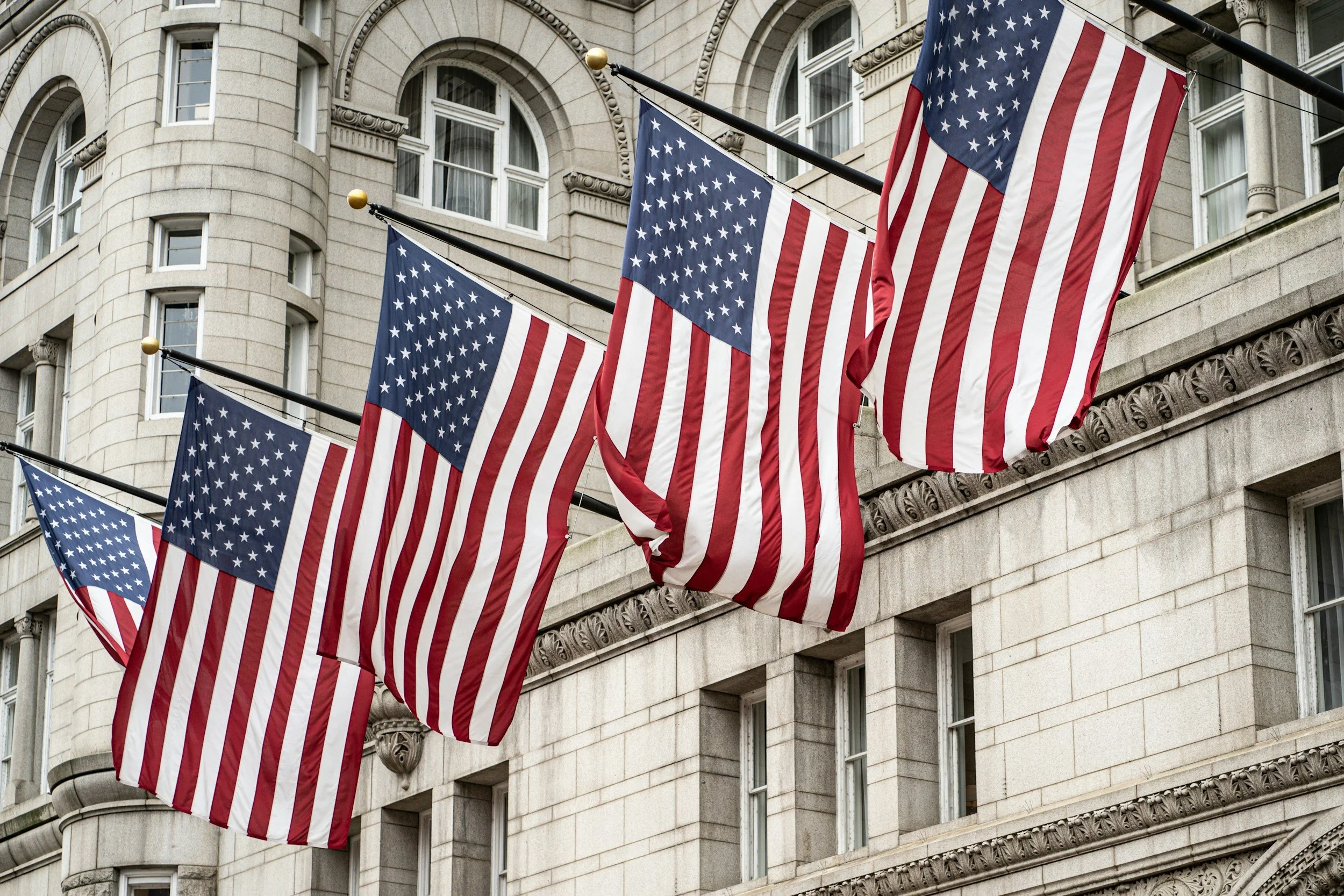 Multiple American flags hanging on flagpoles in front of a large stone building.