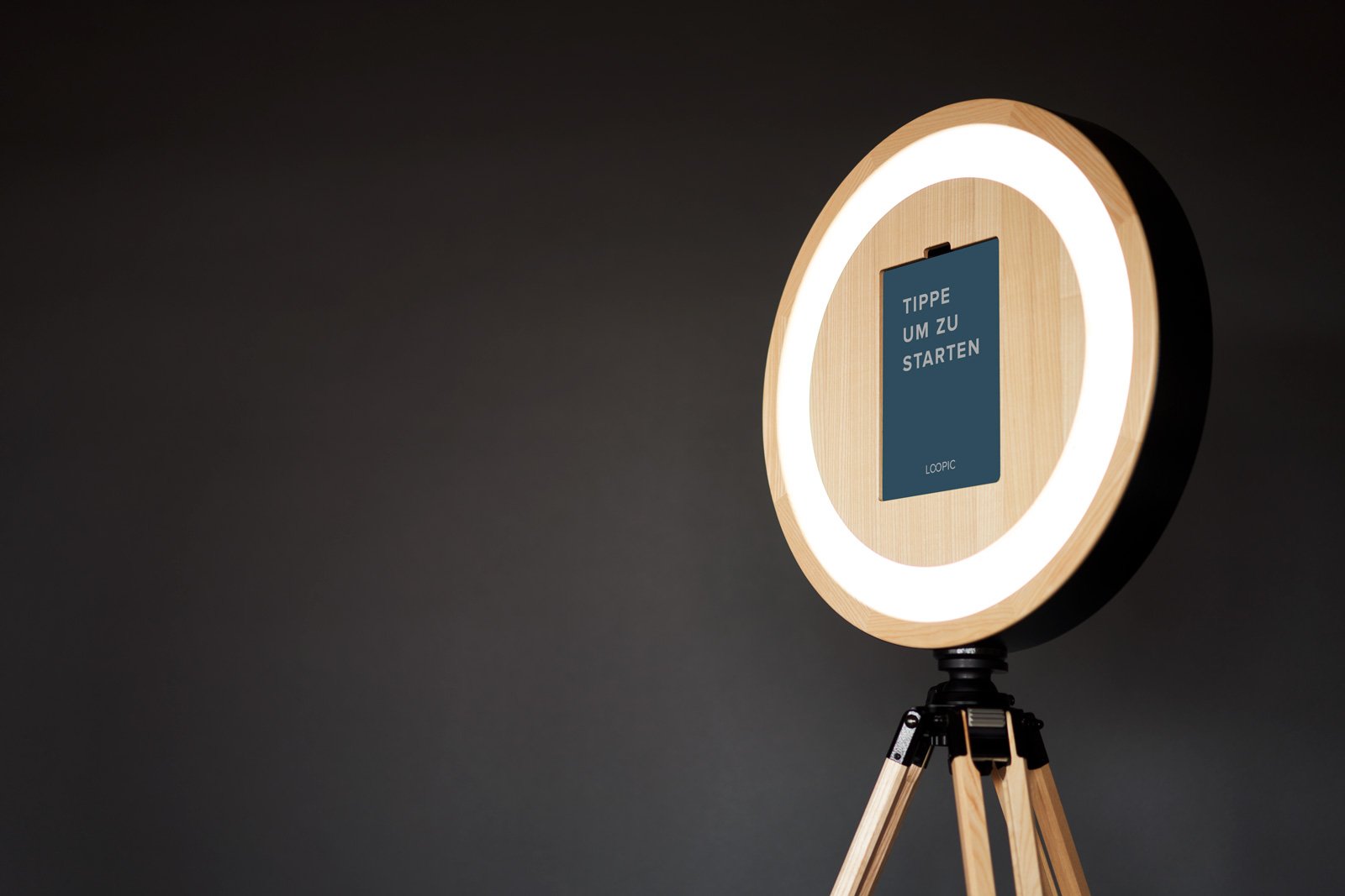 Ein runder Spiegel mit LED-Beleuchtung auf einem Holz-Stativ, im Inneren ein Smartphone und der Text 'TIPPE UM ZU STARTEN'.