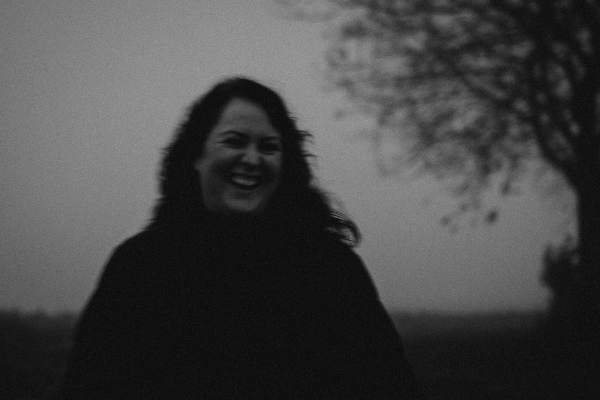 Schwarz-Weiß-Foto einer lachenden Frau im Freien mit einem Baum im Hintergrund.