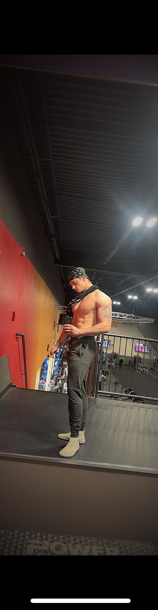 A shirtless man with a bandana and tattoos taking a mirror selfie in a gym, wearing black pants and beige socks.
