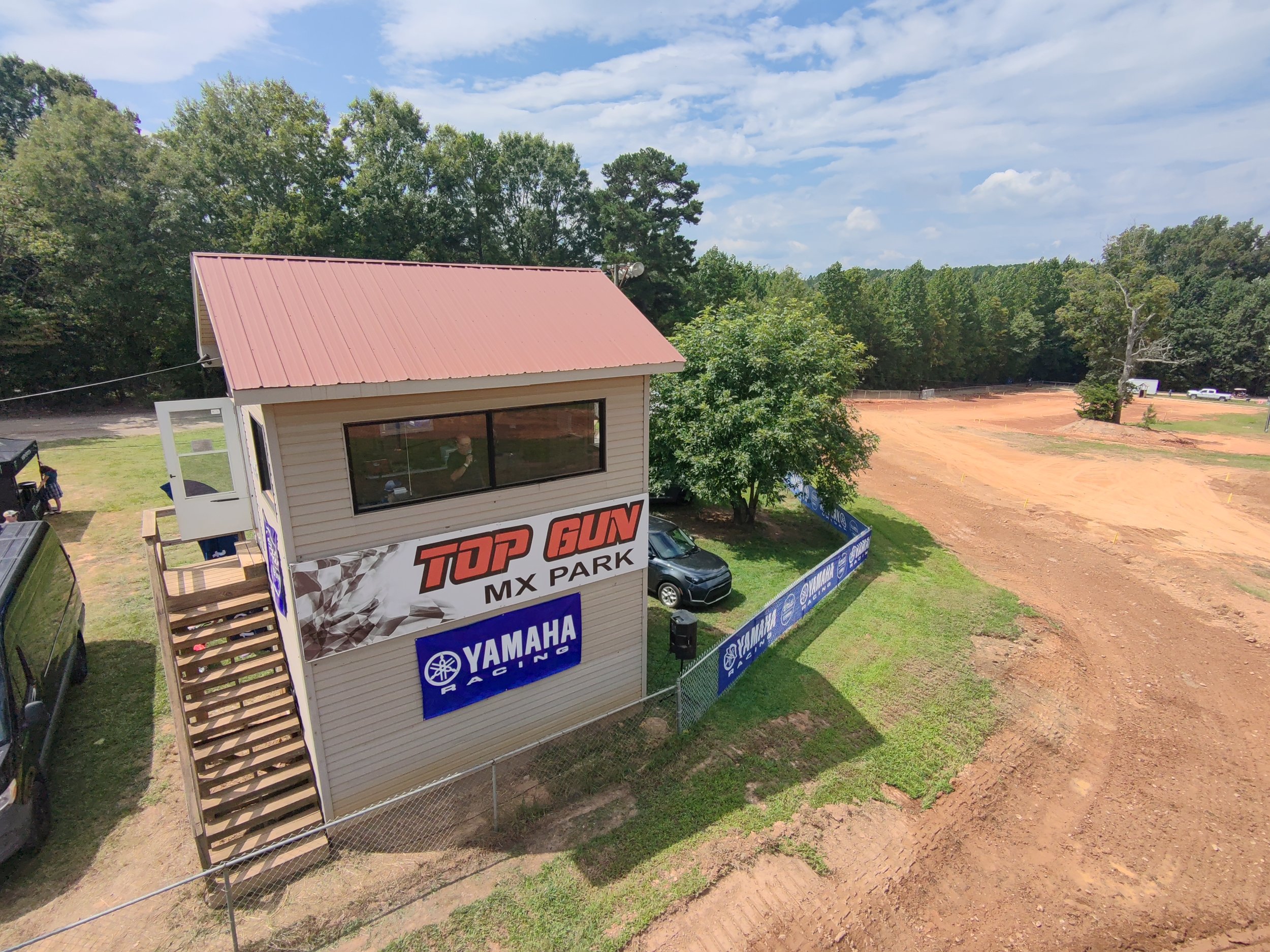 A two-story building with a pink metal roof, featuring a sign that reads 'Top Gun MX Park' and a Yamaha Racing logo. The building has large windows and an outdoor staircase leading to a second-floor entrance. Next to the building is a small parking area with a black car. The area is fenced and surrounded by green trees, with a large dirt track or open field adjacent to the property.