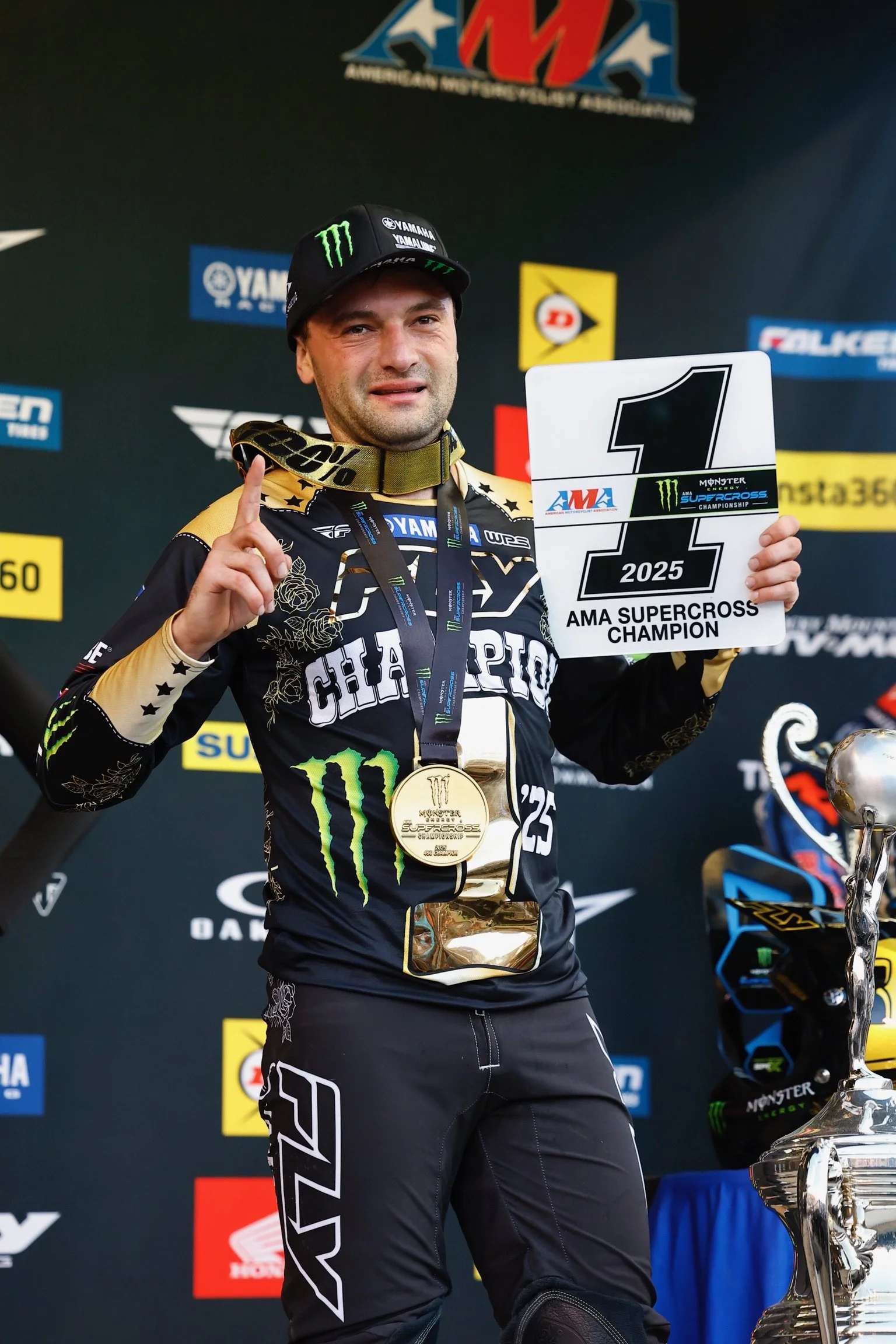 Cooper Webb in racing gear holding a sign that reads '2025 AMA Supercross Champion' and wearing a gold medal. He is standing in front of a backdrop with sponsor logos.