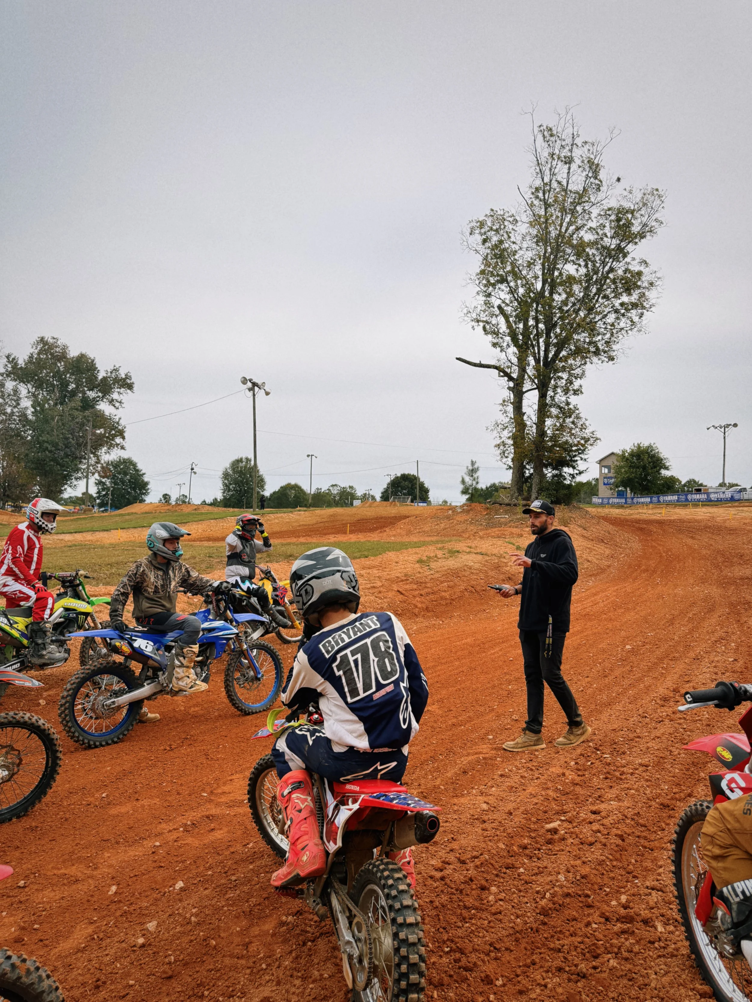 Youth motocross racers gathered around a dirt track, with a coach or instructor providing instructions, on an overcast day with a large tree and some buildings in the background.