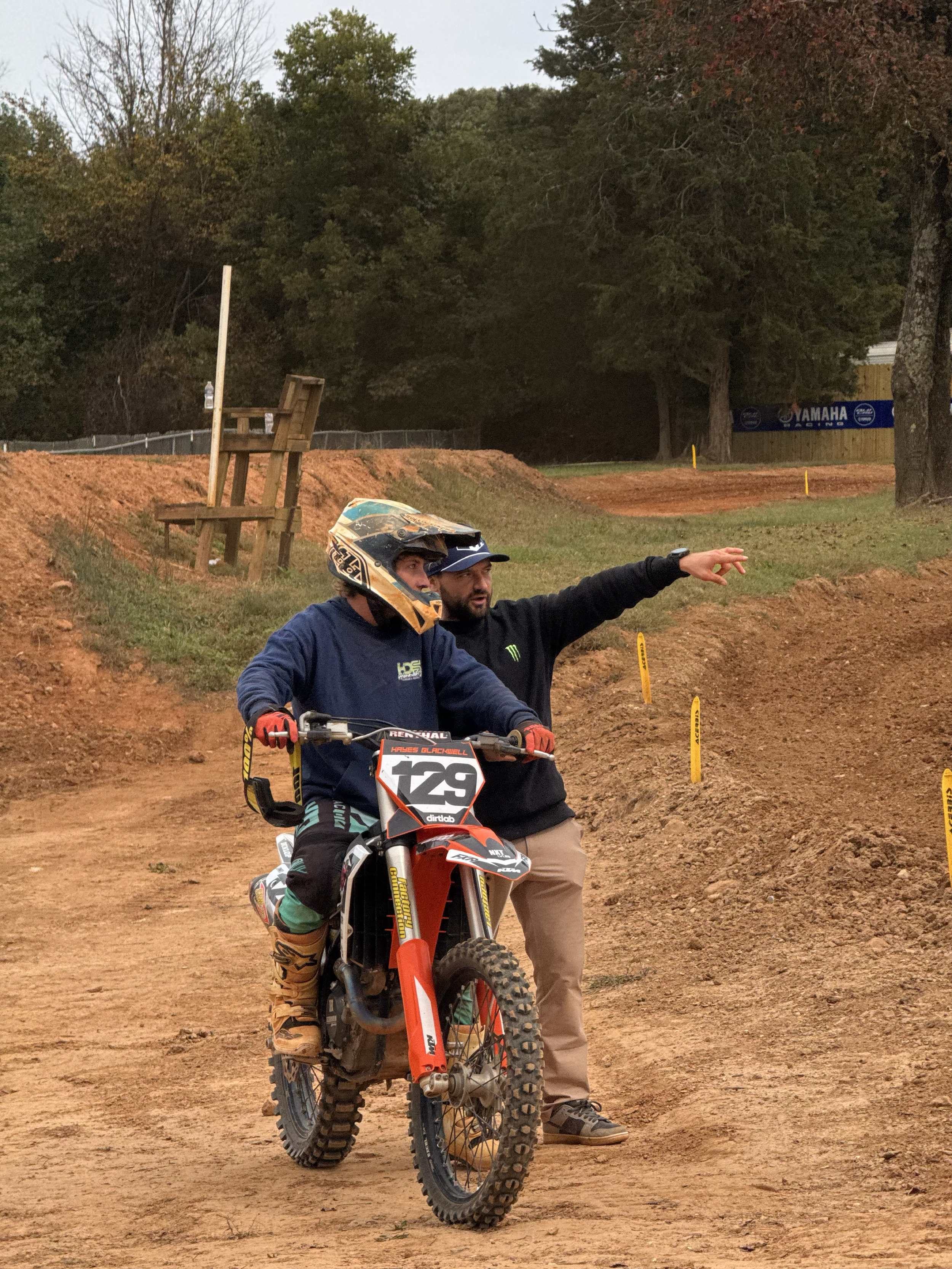 A dirt bike rider wearing a helmet and jeans sitting on a motocross bike, with a man in a black hoodie and beige pants pointing and giving instructions on a dirt motocross track.