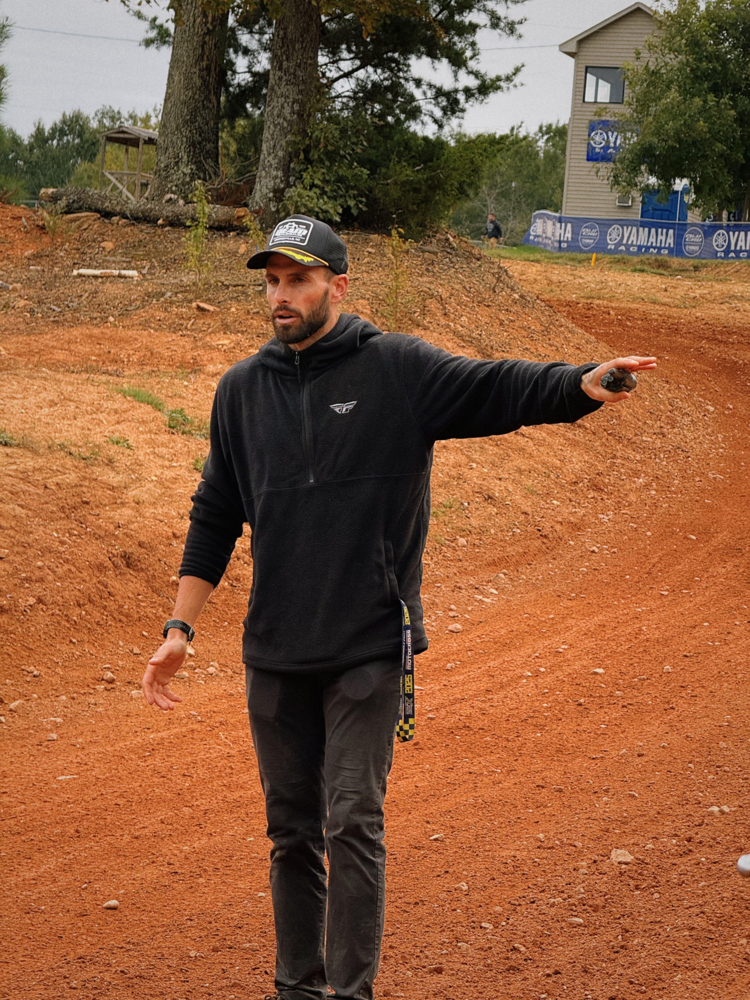 A man standing on a dirt trail, pointing to the right, with trees and a building in the background.