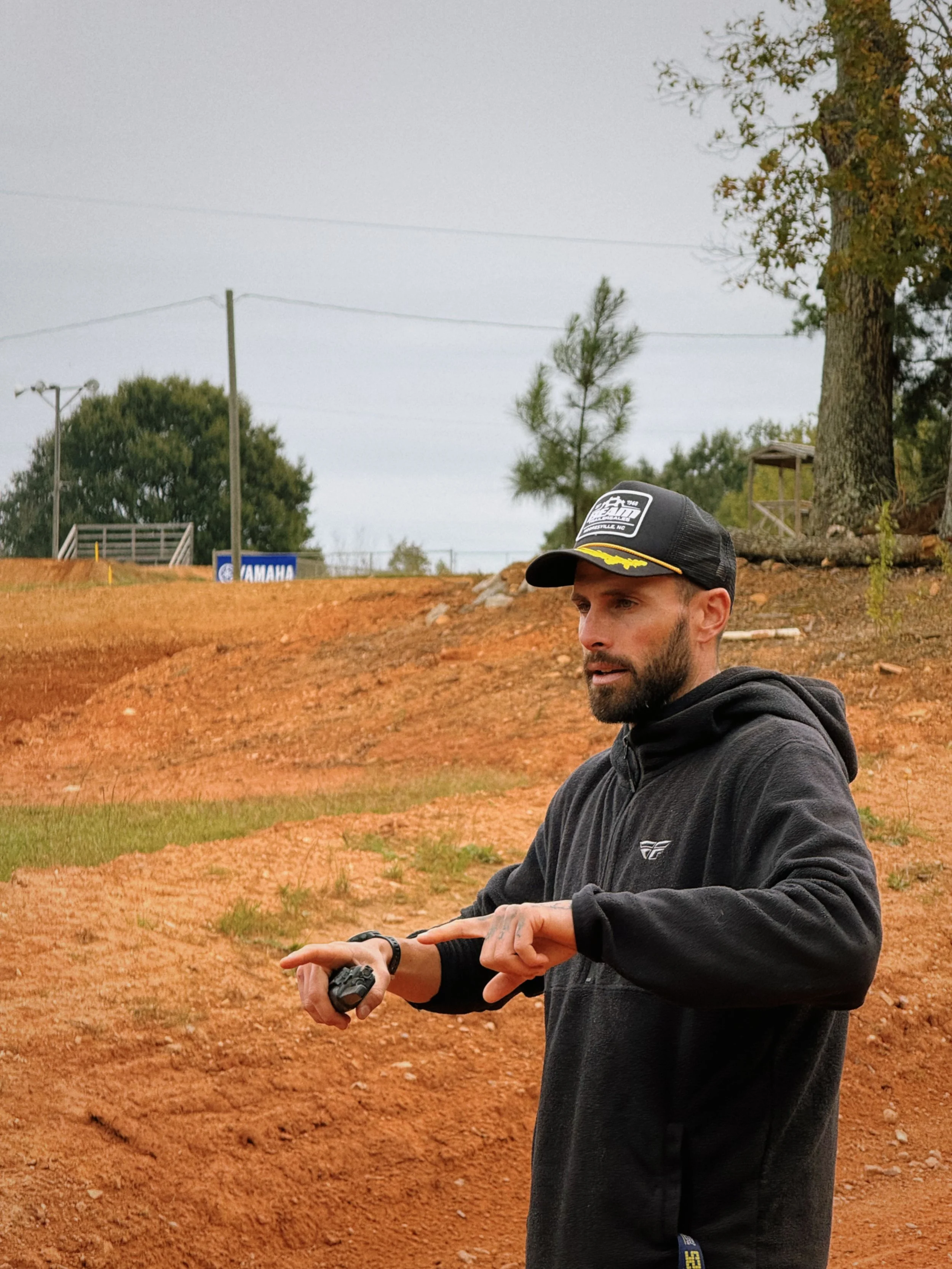A man with a beard wearing a black hoodie and cap, holding a stopwatch, standing outdoors on a dirt track with trees and power lines in the background.
