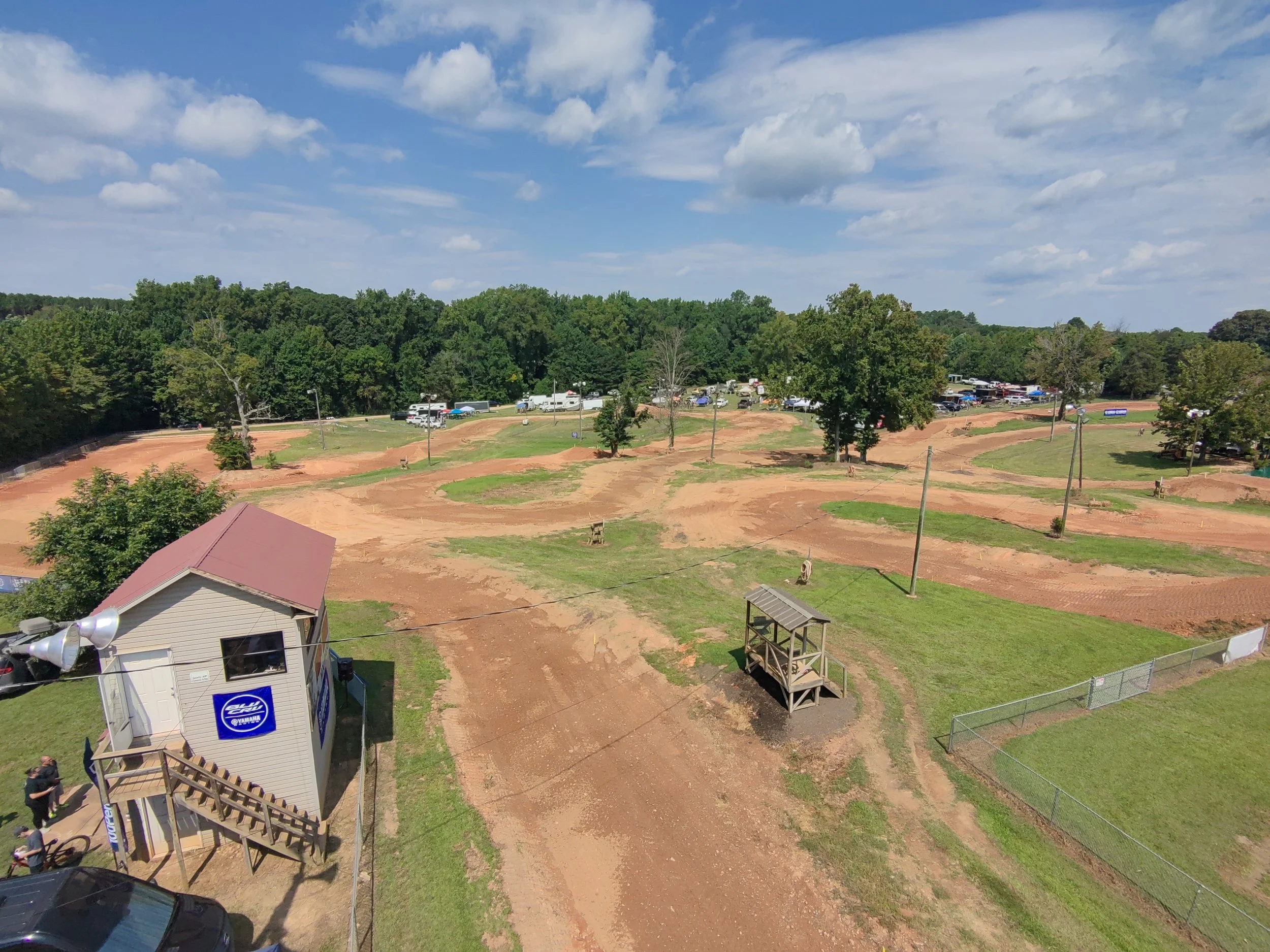 An dirt bike track in a park with winding dirt paths, surrounded by trees, parked vehicles, and a small building with a red roof, under a partly cloudy sky.