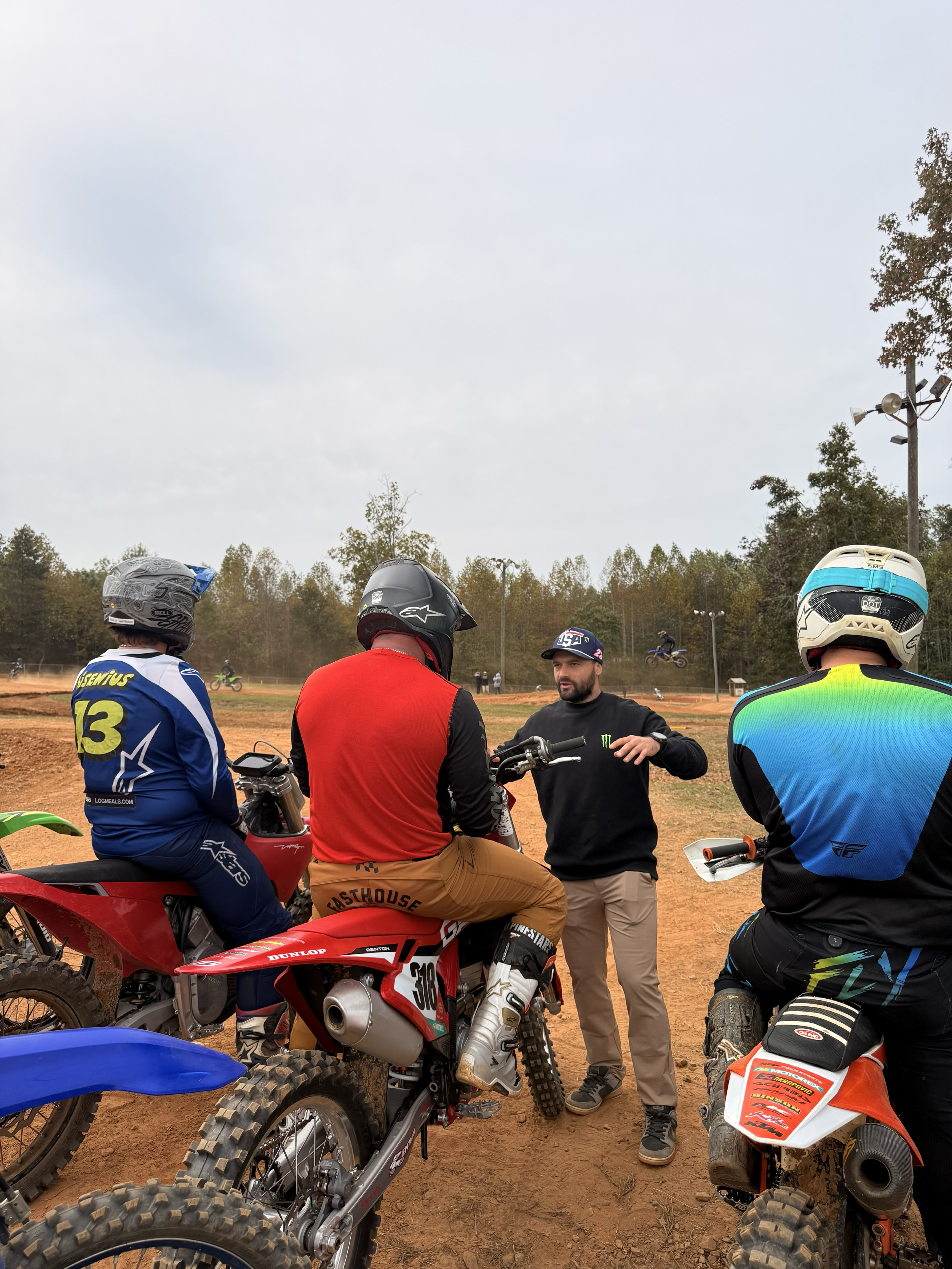 Group of motocross riders in gear and helmets sitting on bikes, listening to a man in casual clothes talking outdoors at a dirt track.