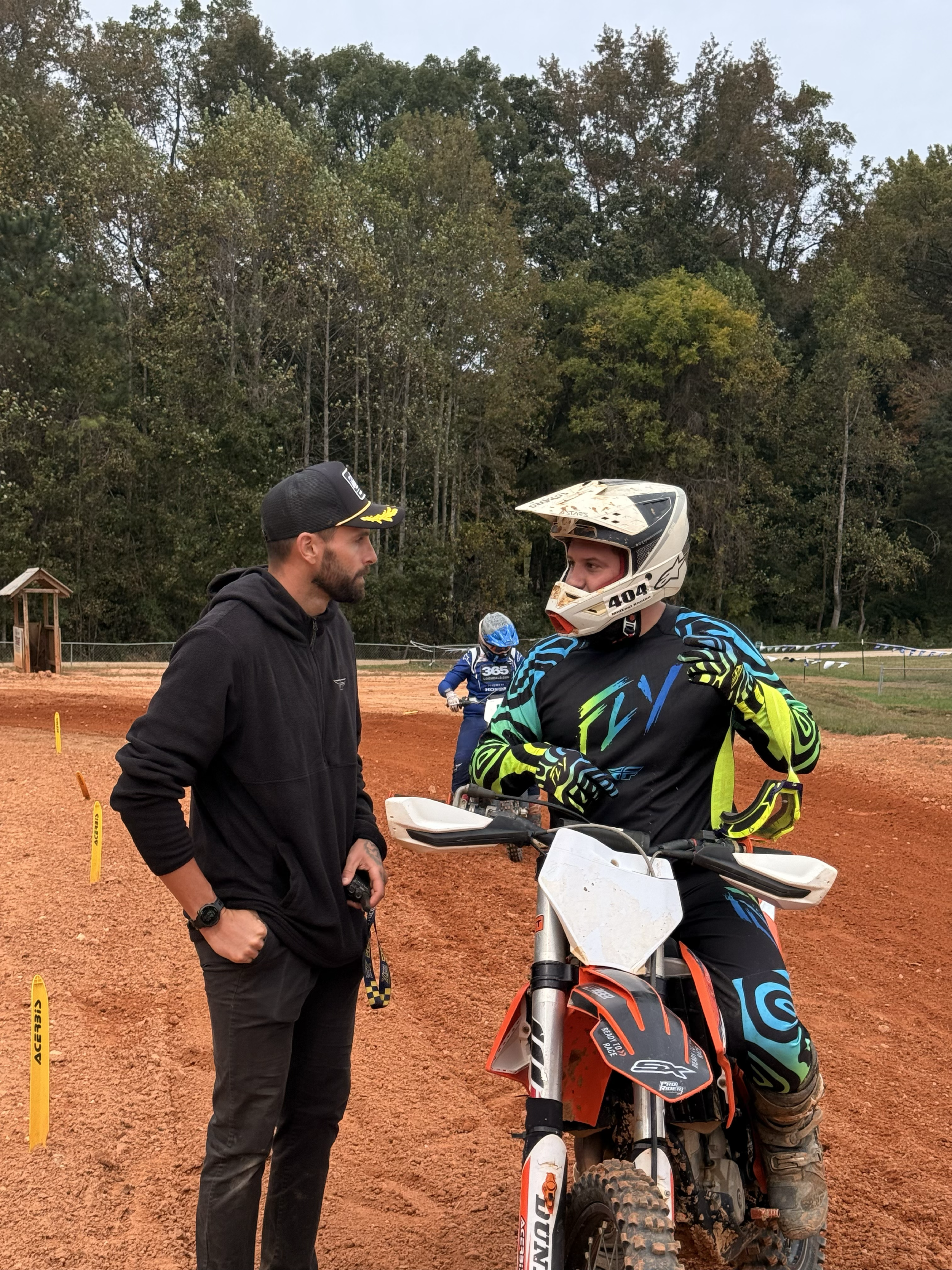 A man in a black hoodie and another man in motocross gear, including a helmet, are having a conversation on a red dirt motocross track. There are trees in the background and a person in blue motocross gear riding a dirt bike in the distance.