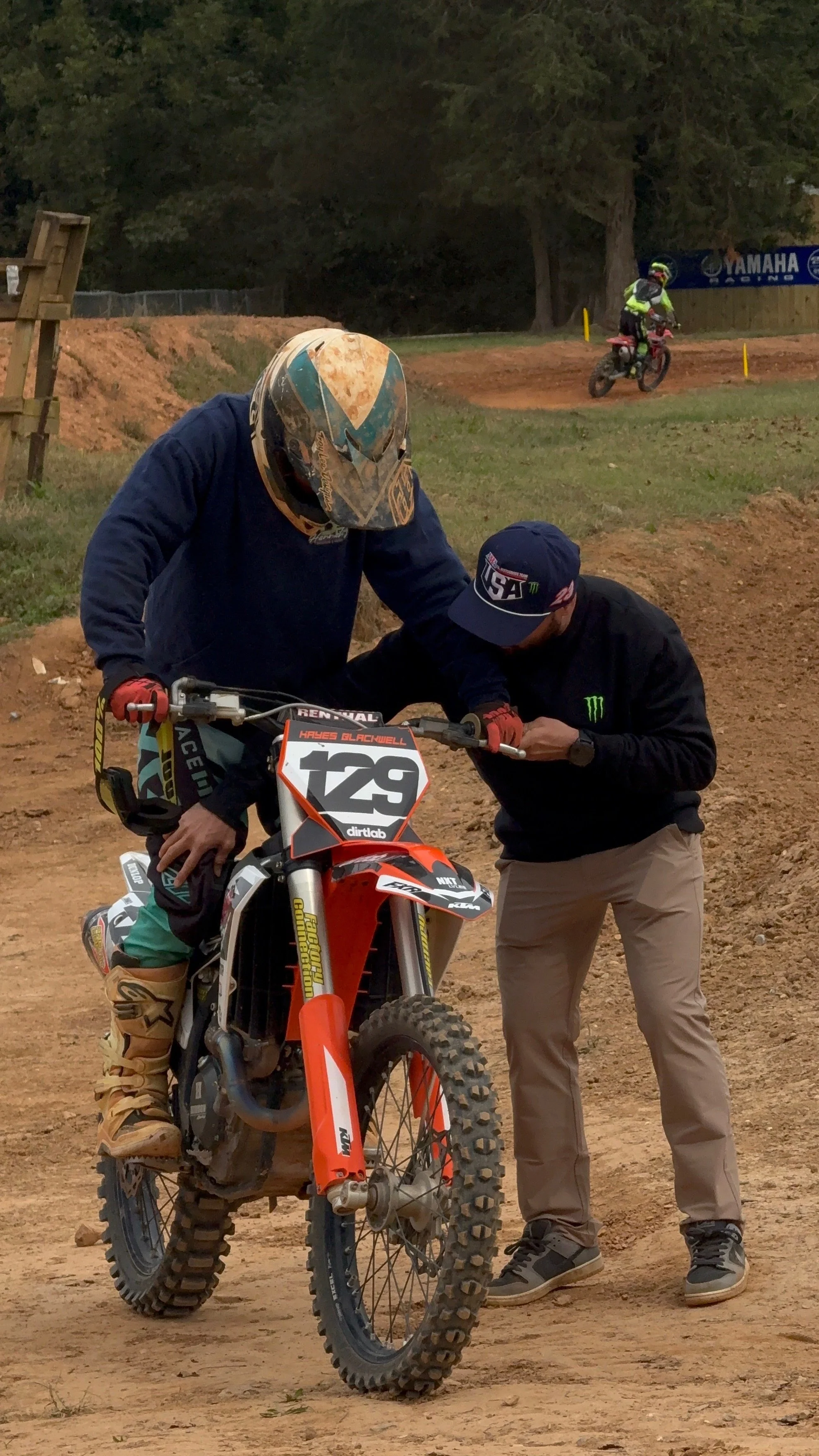 A person in a blue jacket and a helmet assisting a rider on an orange dirt bike with the number 129 on a dirt track. In the background, another rider is seen on a motorcycle on the track.