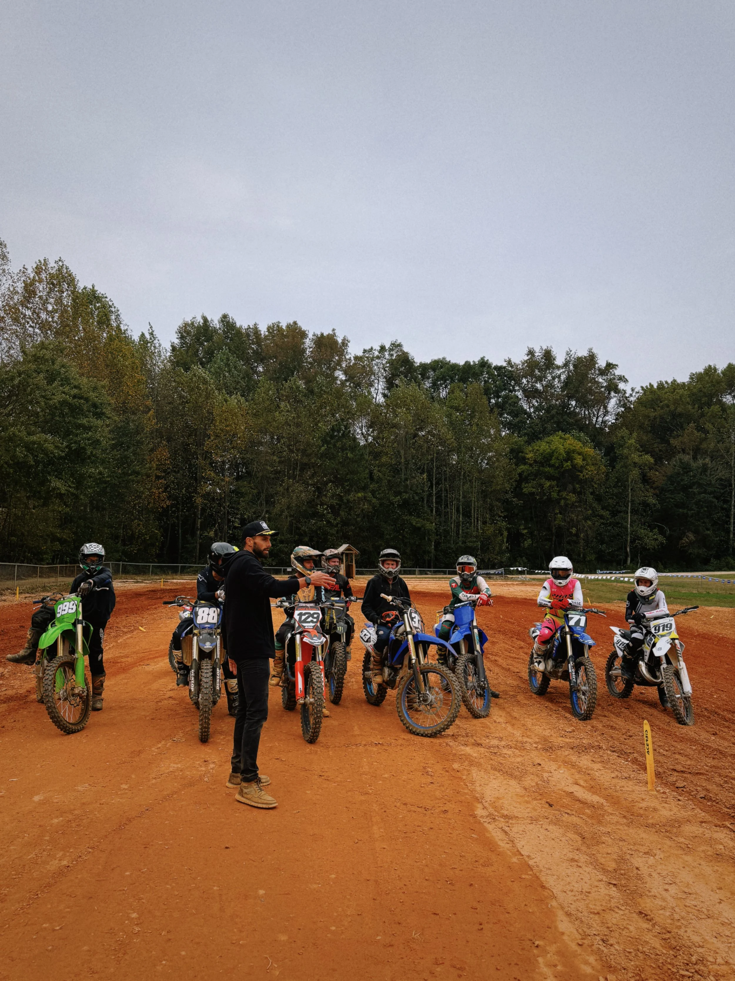 A group of eight motocross riders with helmets and racing gear on dirt bikes, with a person in black standing in front of them, on a dirt track surrounded by trees.