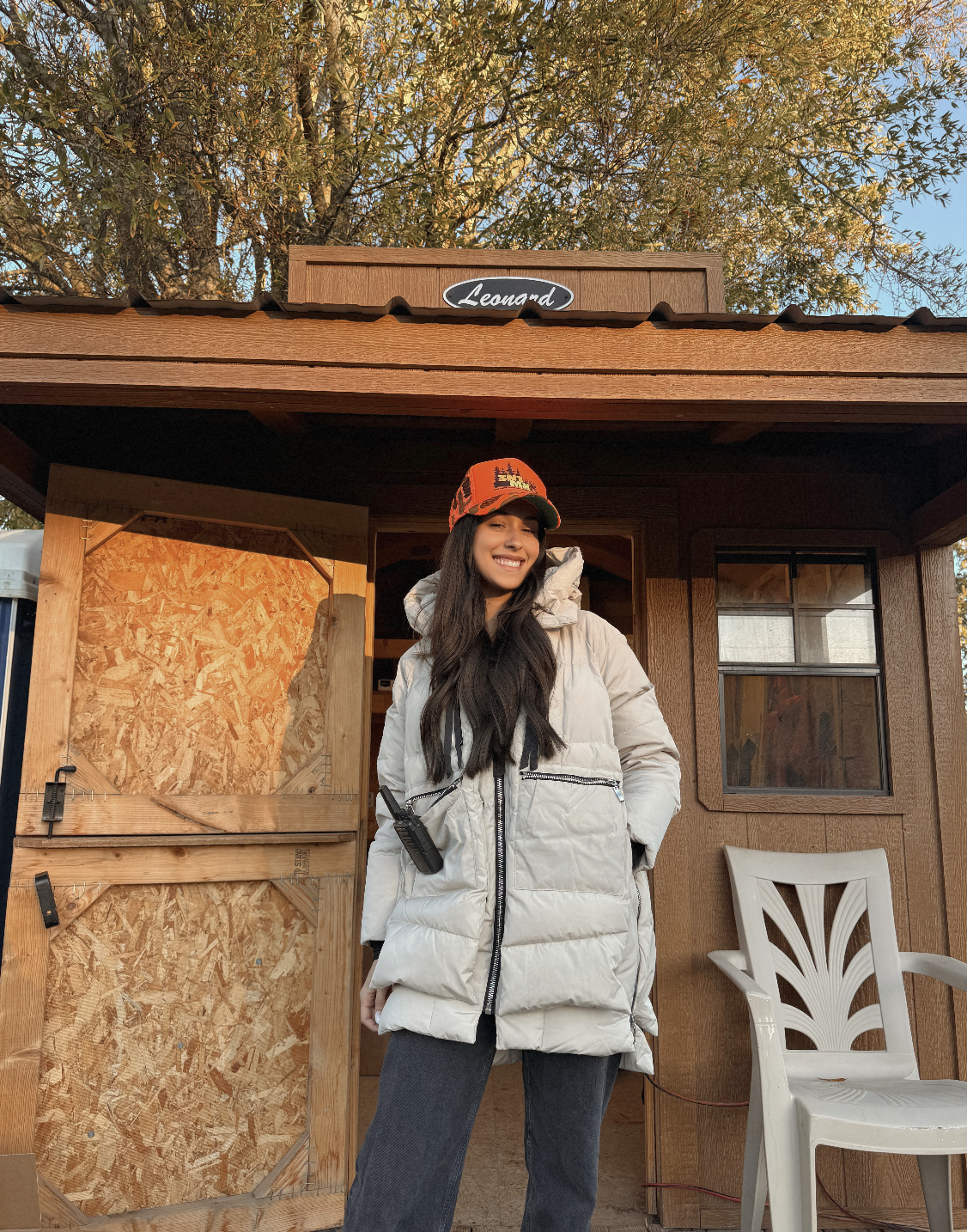 A woman in a beige puffer coat and orange cap standing outside a small wooden structure with a sign labeled 'Leonard' on the roof, autumn leaves and a tree in the background, smiling.