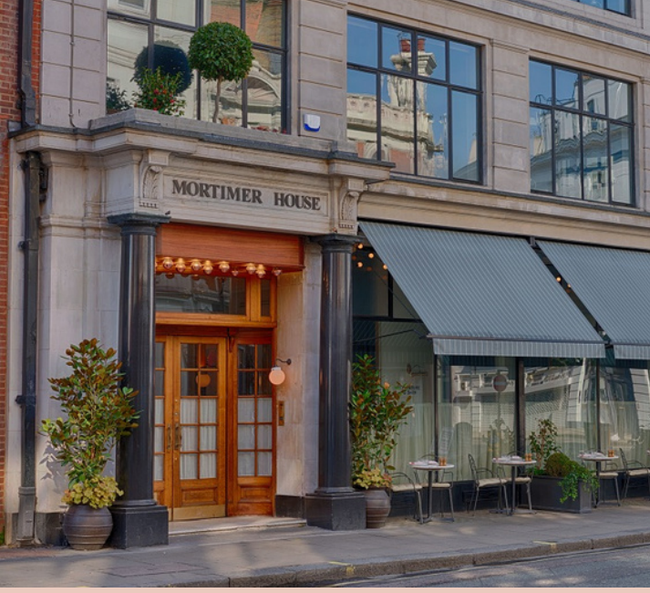 The exterior of the Mortimer House building featuring a wooden entrance with glass doors, black columns, and potted plants on either side, with dining tables and chairs outside covered by awnings.