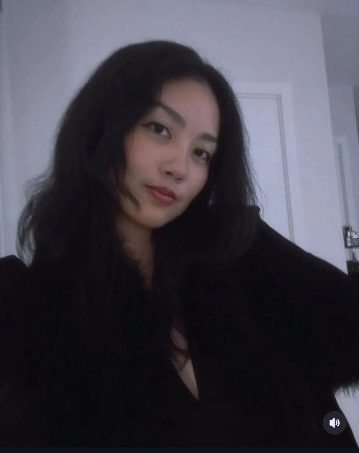 A young woman with long dark hair posing for a selfie in front of a white door, wearing a black top, with a neutral expression.