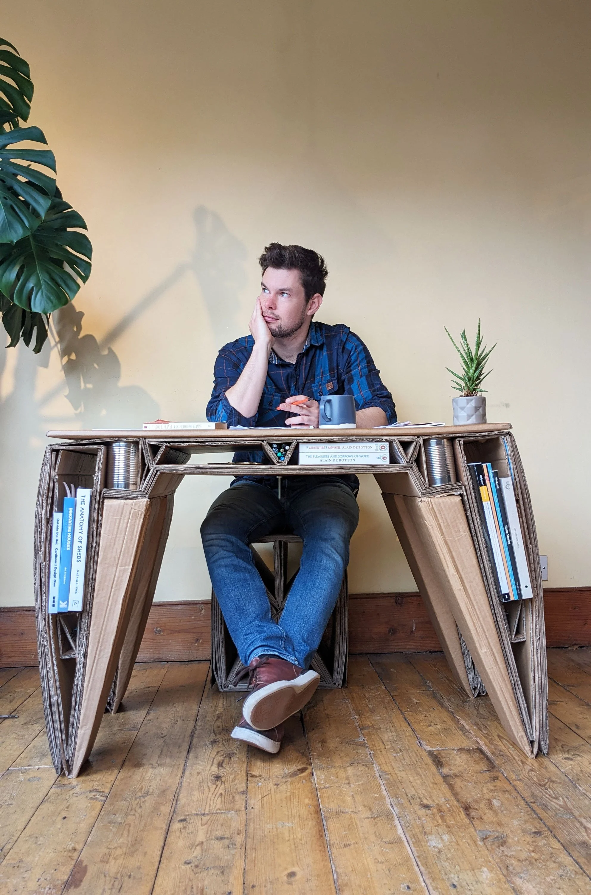 A young man with short, dark hair sits at a desk made of cardboard, with a thoughtful expression, resting his face on his hand, and a phone in the other hand. The desk is filled with books and a mug, with a large green plant and a smaller succulent on the desk. The room has wooden floors and a plain beige wall.
