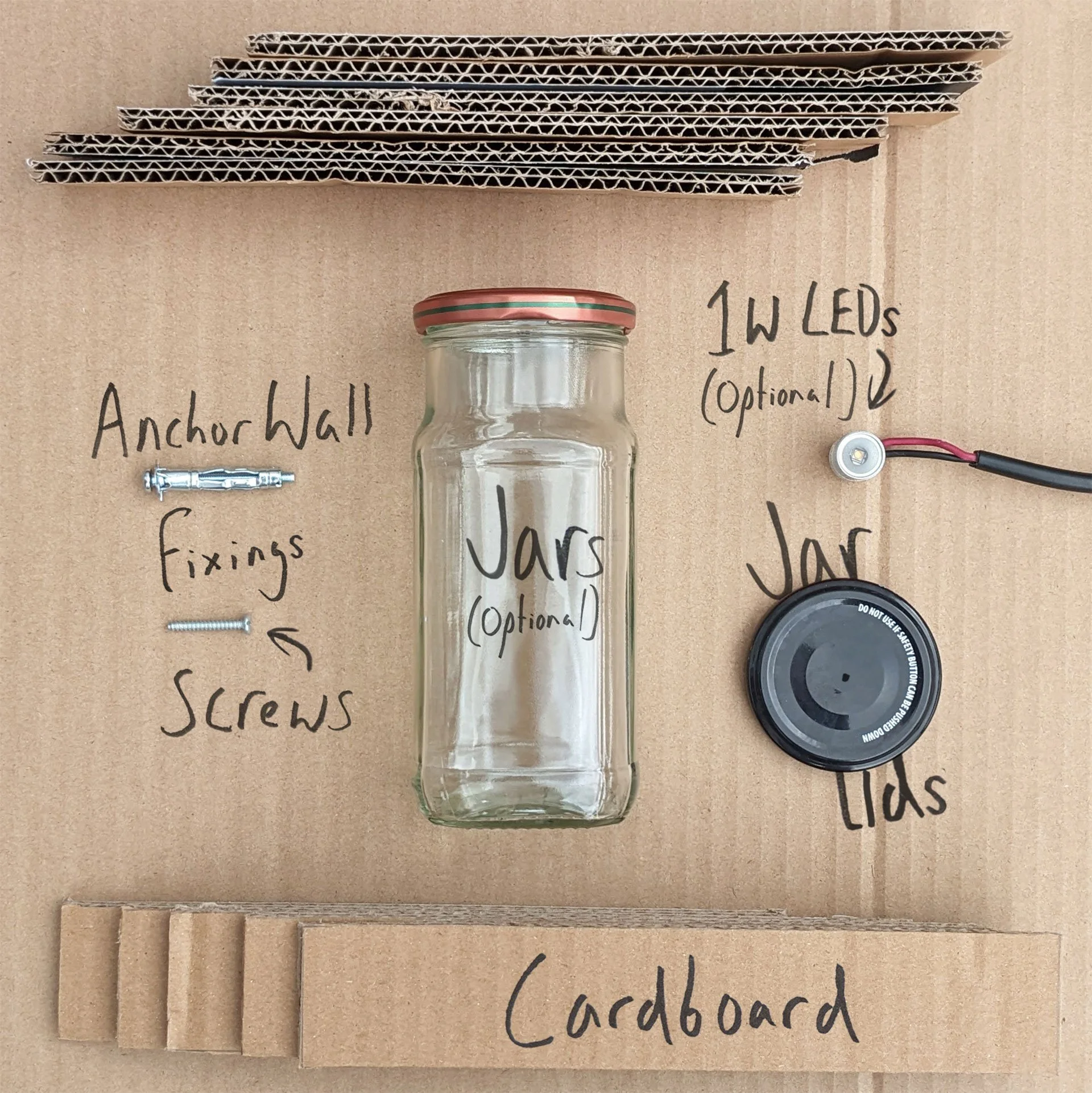 Flat cardboard surface with hand-written notes, a glass jar, and various hardware components for a project, including pieces labeled as 'anchor wall,' 'fixings,' 'screws,' 'washer,' and 'grabbord'.