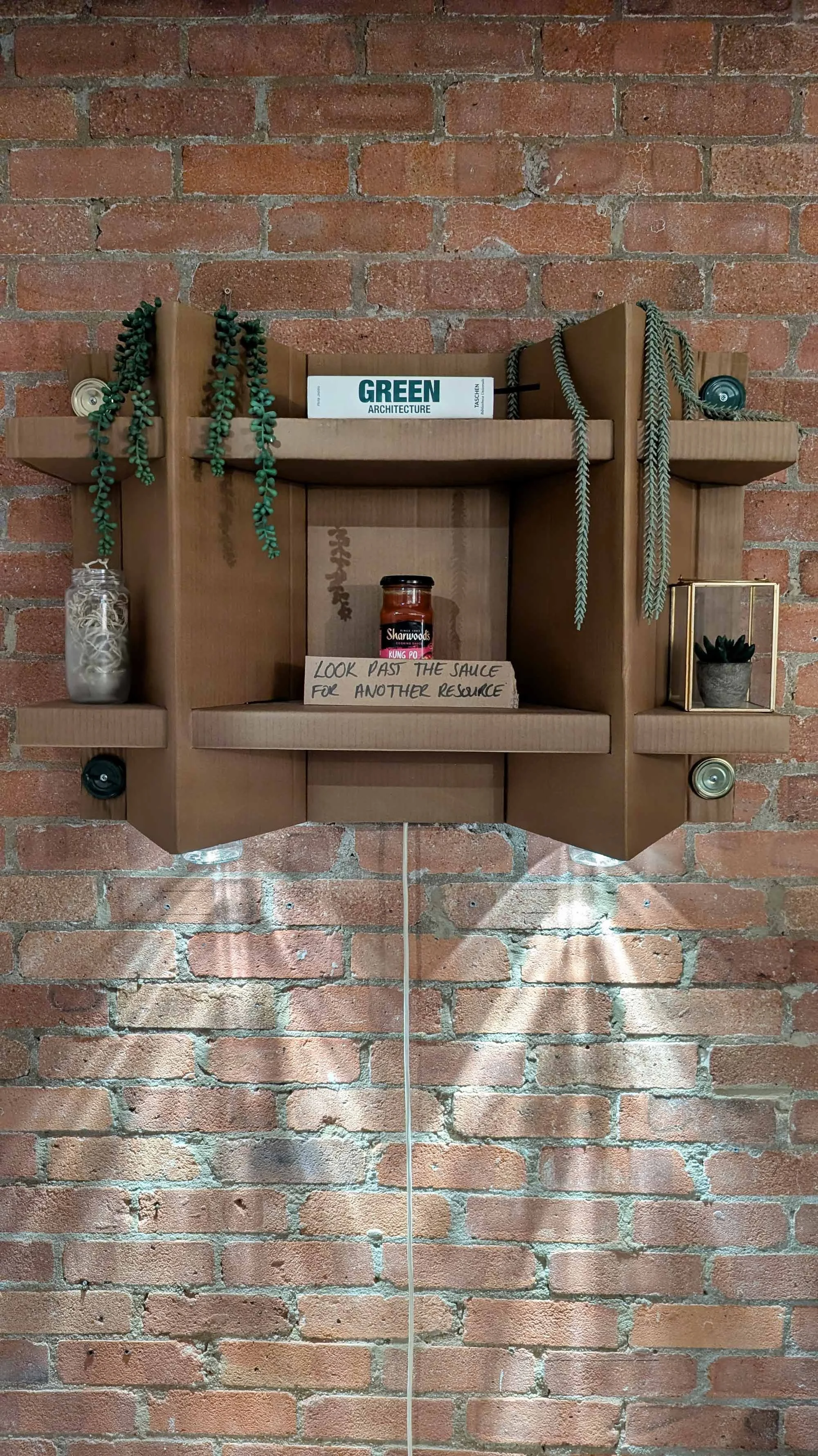 A shelving unit with lighting made from upcycled cardboard and glowing glass jars mounted on a brick wall, decorated with pot plants, an architecture book, a jar of sauce, with a handwritten note that says, 'Look past the sauce for another resource'.