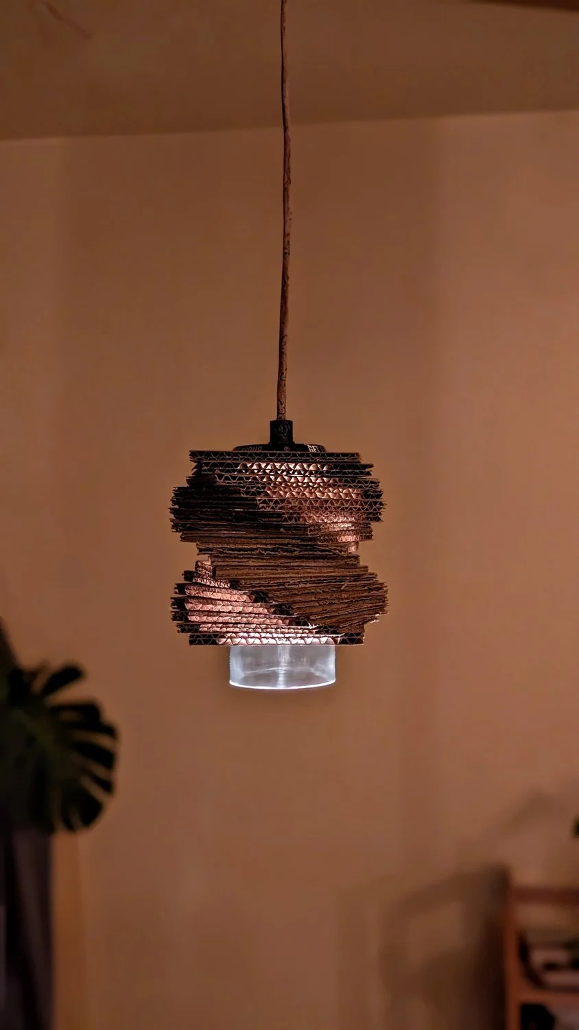 A pendant light made of layered, wavy cardboard pieces hanging from a cord in a room with beige walls.