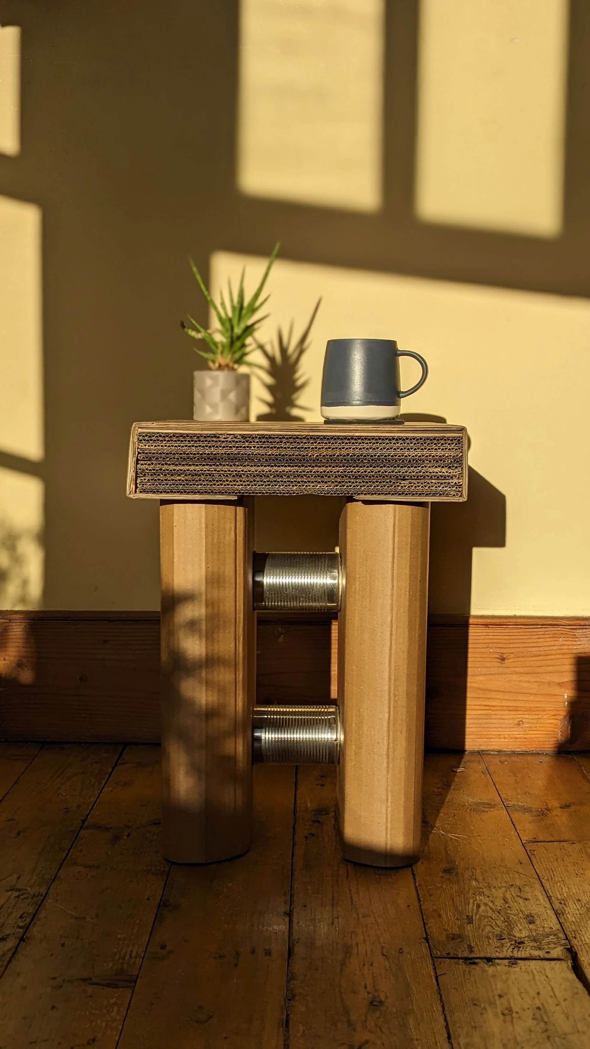 A small table made of upcycled cardboard and metal cans, with a potted plant and a mug placed on it, casting shadows on the yellow wall behind.