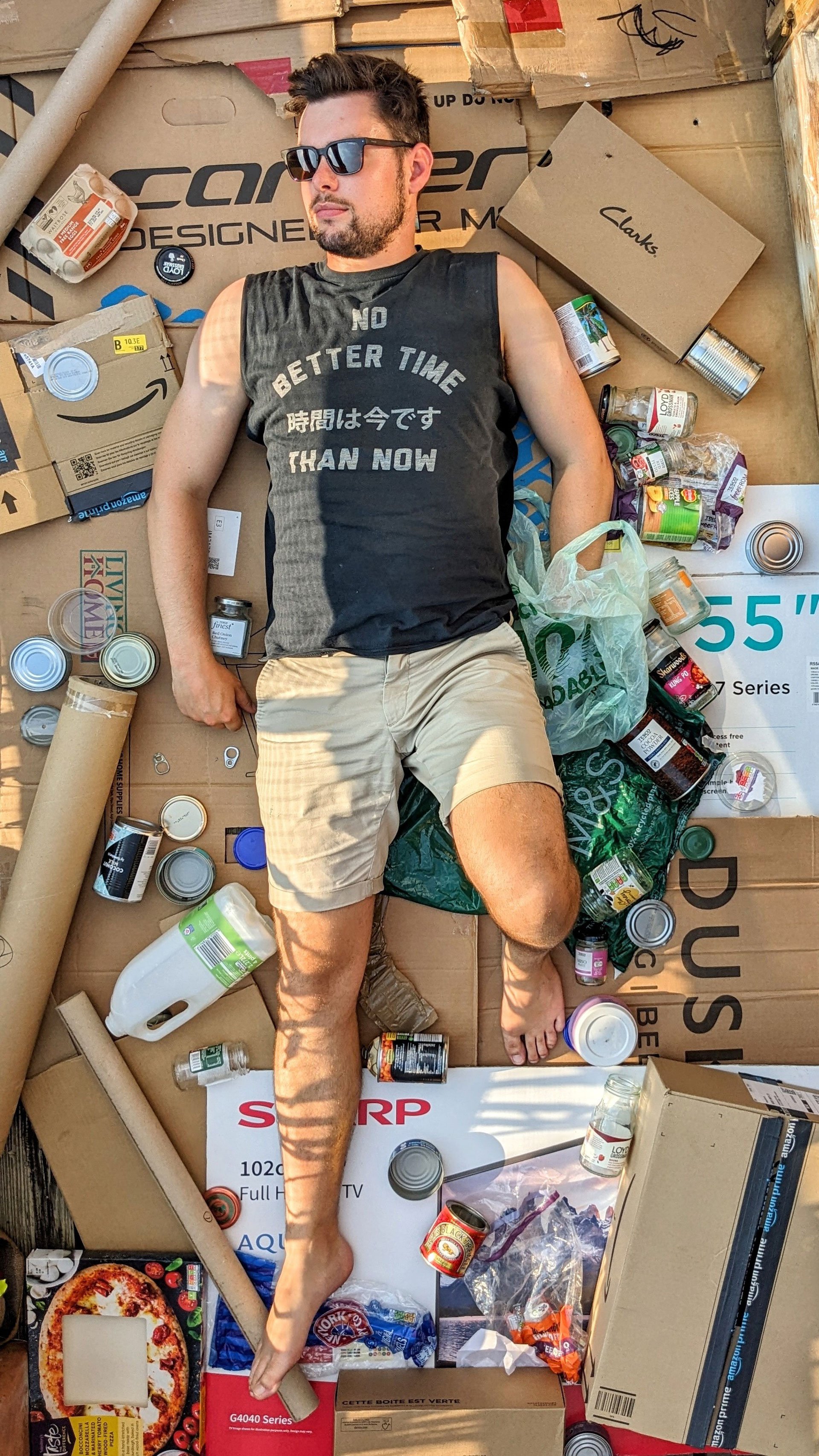 A man lying on the ground surrounded by discarded food cans, boxes, and trash, wearing sunglasses, a black sleeveless shirt, and shorts.