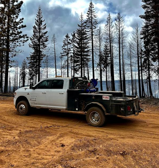A white and black utility truck parked on a dirt road in a forested area with tall, sparse pine trees and a cloudy sky in the background.