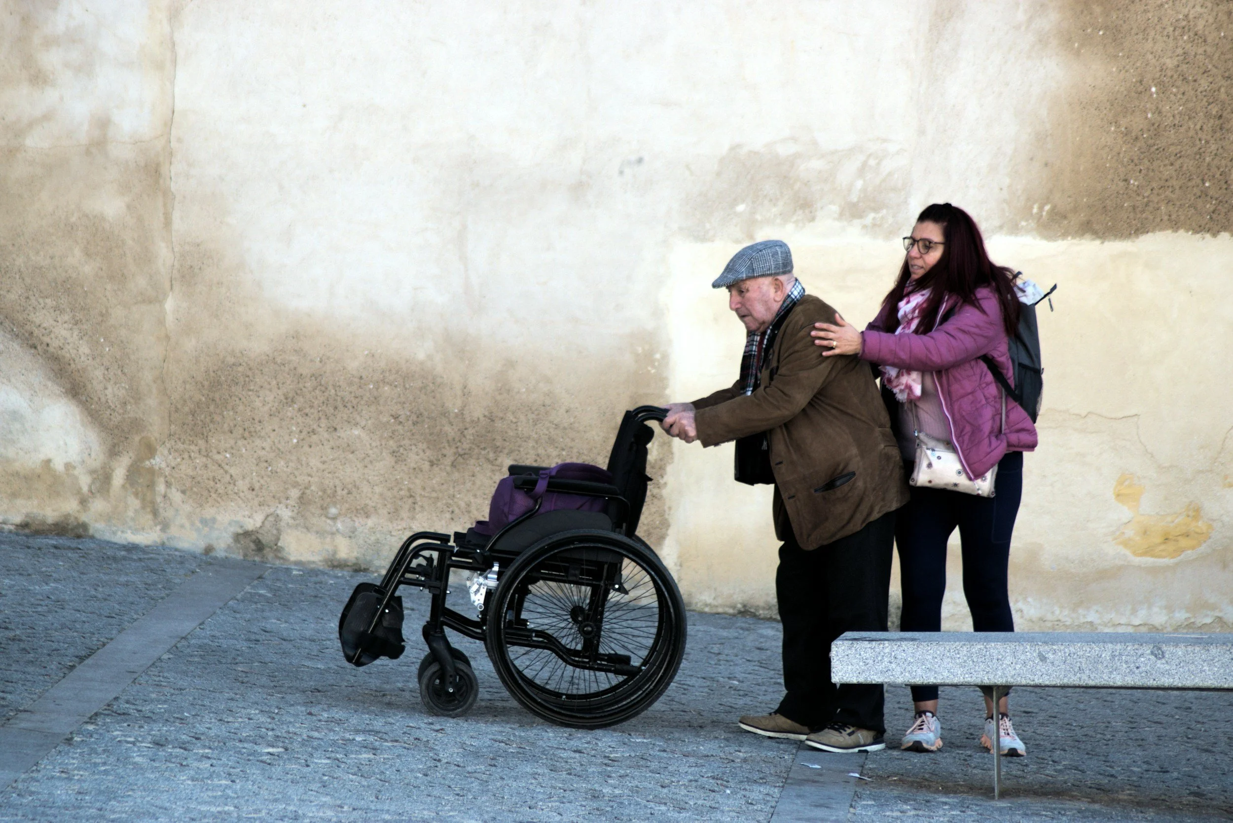A young woman helping an elderly man in a wheelchair walk along a sidewalk against a textured wall. The woman is wearing a pink jacket, glasses, and has a backpack. The man is wearing a brown jacket, cap, and has a scarf.